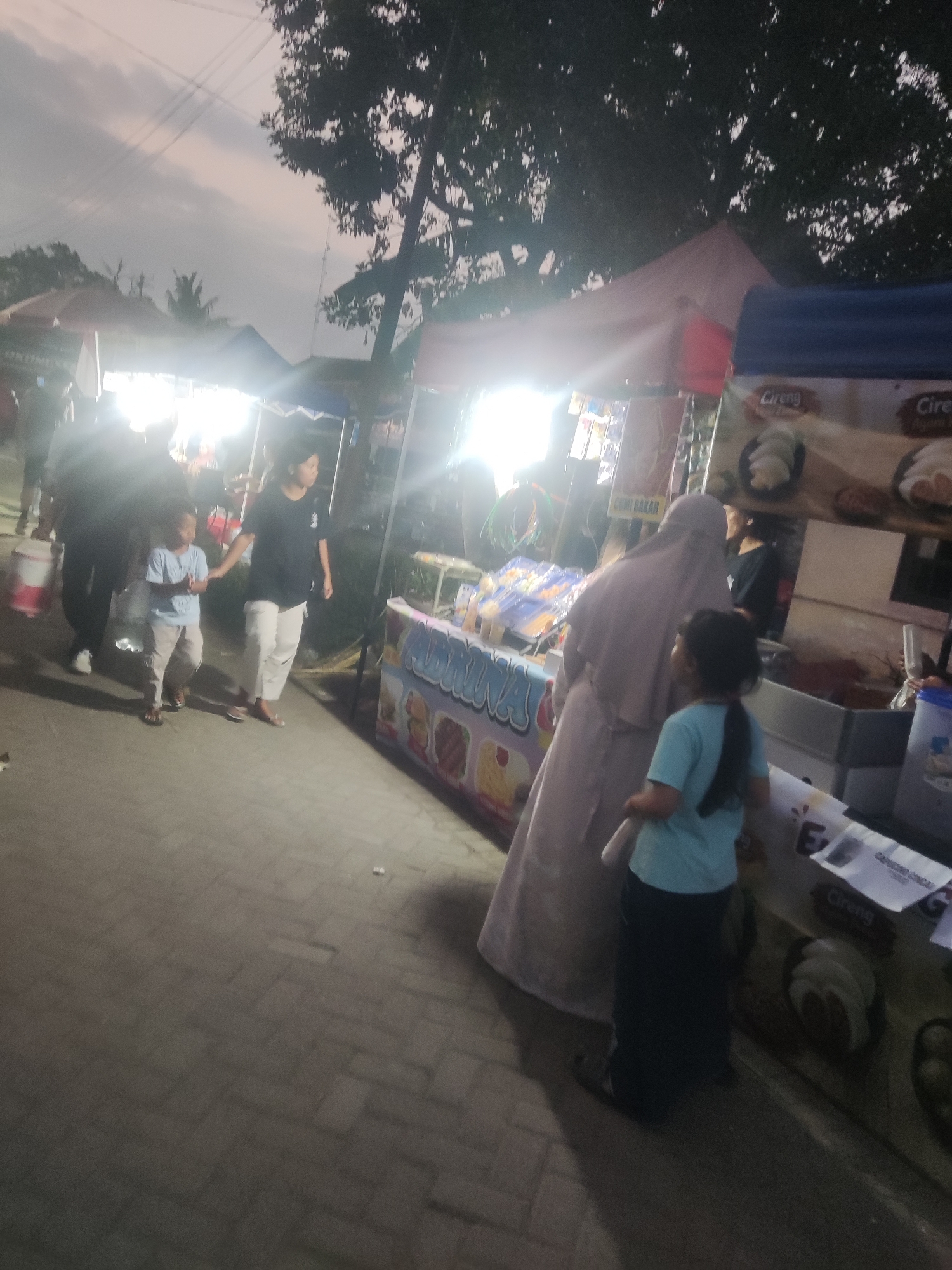 A daytime, medium shot captures an outdoor marketplace scene at dusk, with several food stalls and pedestrians. The foreground shows a paved walkway where two individuals are walking. To the left, a young boy in a light blue t-shirt and light-colored pants, accompanied by a woman in a black t-shirt and white pants, walks away from the camera.
The middle ground is occupied by various food stalls. One stall on the right displays signs with "Cireng" written on them, and images of food items. A person in a light purple hijab and a child in a blue t-shirt are visible near this stall. Another stall to the left, brighter due to illumination, has items displayed and appears to be selling food.
The background reveals more stalls, some with illuminated signs and tents. Trees with dark foliage are visible overhead, along with power lines crisscrossing the sky. The overall ambiance suggests a lively local market. The presence of "Cireng" on the signage points to a location in Indonesia, and the context of an outdoor market aligns with observations from Trihanggo, Indonesia.
