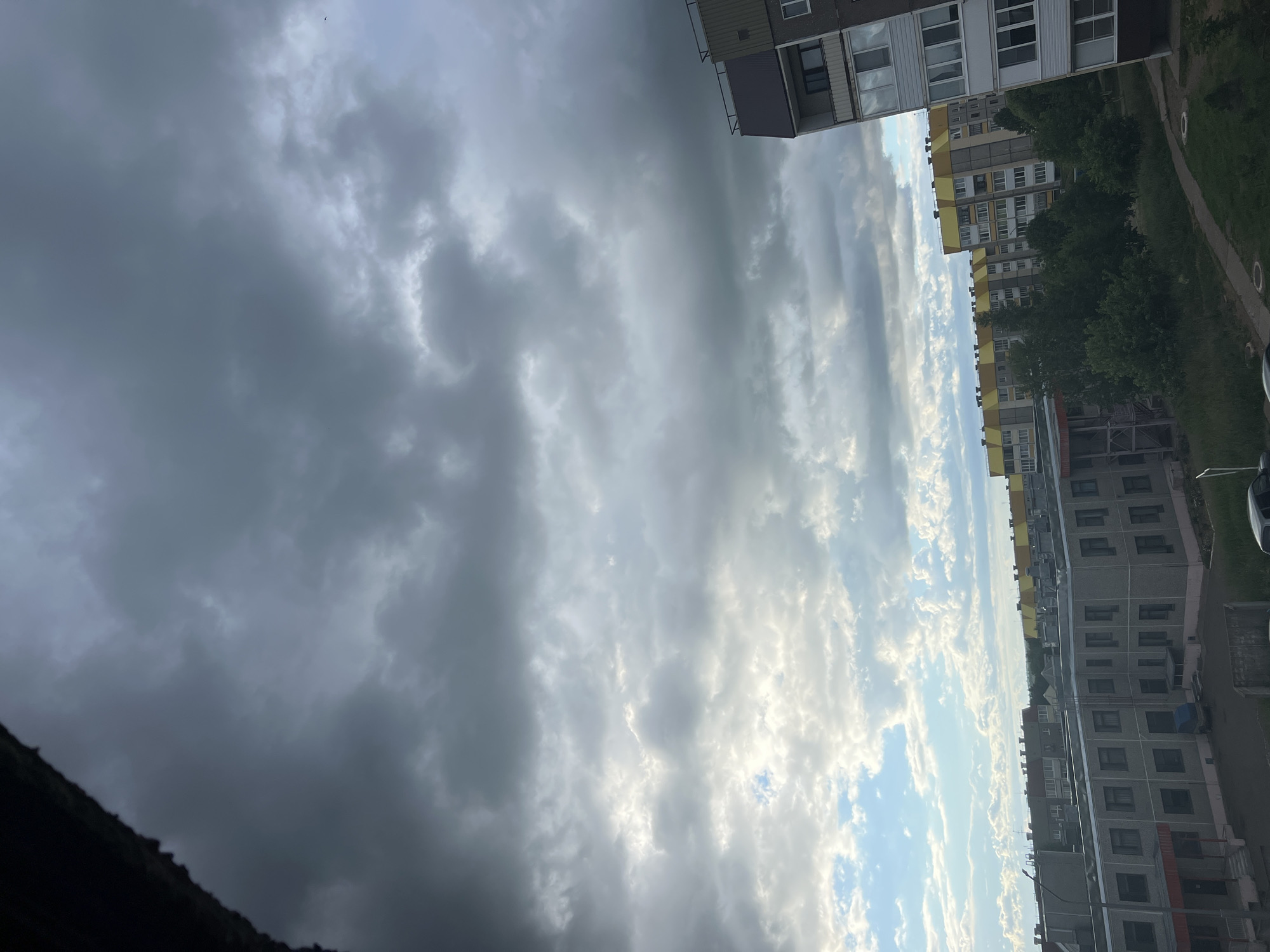 The media file depicts an outdoor scene with apartment buildings and a cloudy sky. The buildings appear to be multi-story residential structures, with visible windows and balconies. The sky is filled with a mix of dark, heavy clouds and lighter, more scattered clouds, suggesting a variable or potentially stormy weather condition. The lighting indicates it is daytime, possibly late afternoon or evening, given the dramatic cloud formations and the angle of light.
The scene is set in an urban or suburban environment, as evidenced by the presence of the apartment complexes and what appears to be a pathway or road to the right of the buildings, bordered by grass. Trees are visible between the buildings, adding a touch of greenery to the scene. The perspective is from a slightly elevated position, possibly a balcony or window of another building, looking out over the complex.
No people are clearly visible in the image, and the focus is on the architecture and the sky. The overall mood evoked by the image is somewhat dramatic due to the strong contrast between the dark and light clouds, hinting at the possibility of changing weather. The location context of Марковский, Russia, suggests the geographical setting. There is no visible text in the image.