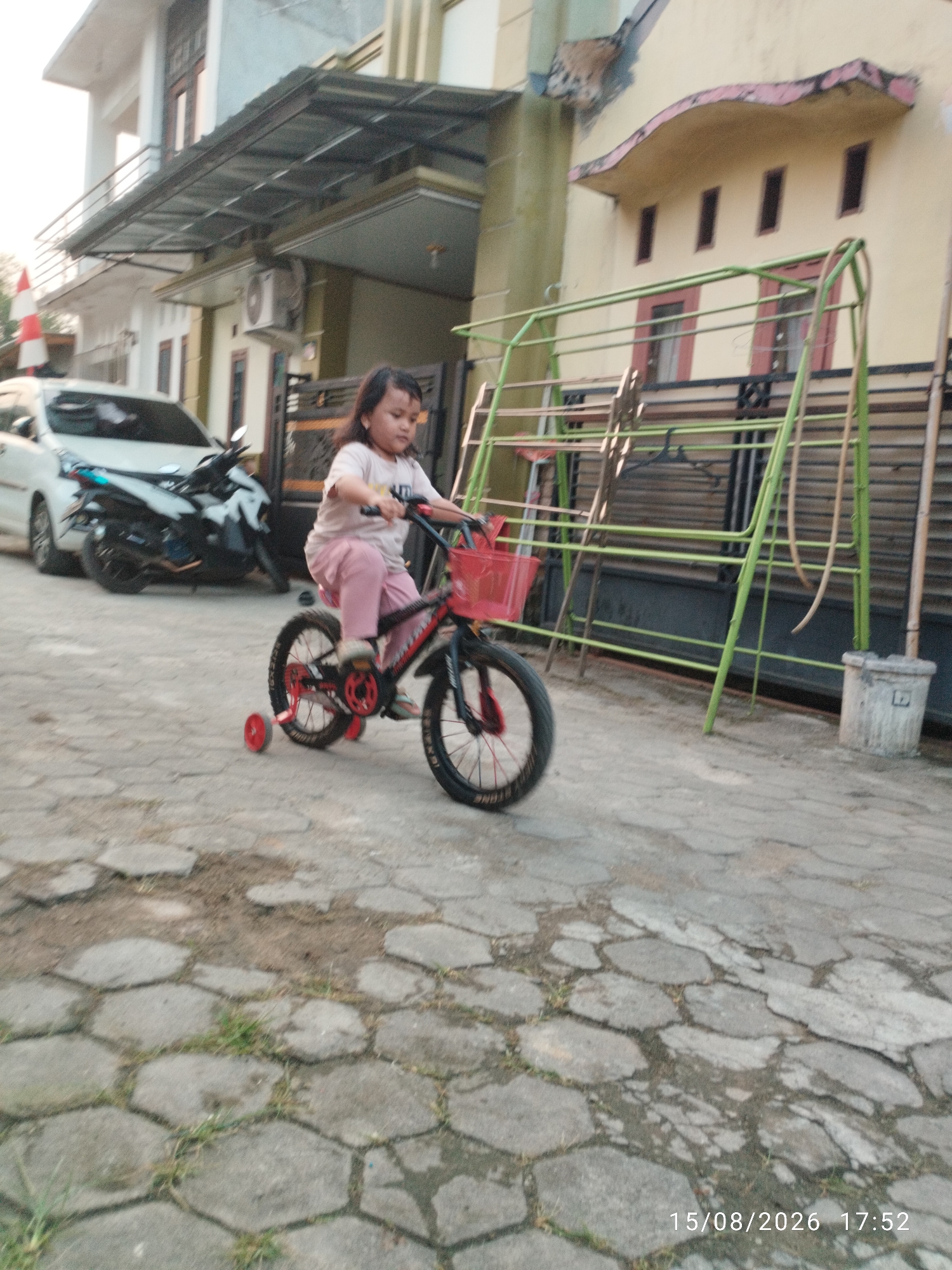 A young girl, appearing to be of Southeast Asian descent, is riding a small black and red bicycle with training wheels on a paved hexagonal stone surface. She is wearing a light pink t-shirt and pink pants. Her bicycle has a red basket attached to the handlebars. In the background, a white car and a dark motorcycle are parked on the street. Behind the parked vehicles, there is a two-story house with a metal awning. To the right of the house, there is a green metal swing set. The scene appears to be in a residential area in Bandar Lampung, Indonesia, as indicated by the architectural style and the timestamp "15/08/2026 17:52" which suggests a Southeast Asian date format.
