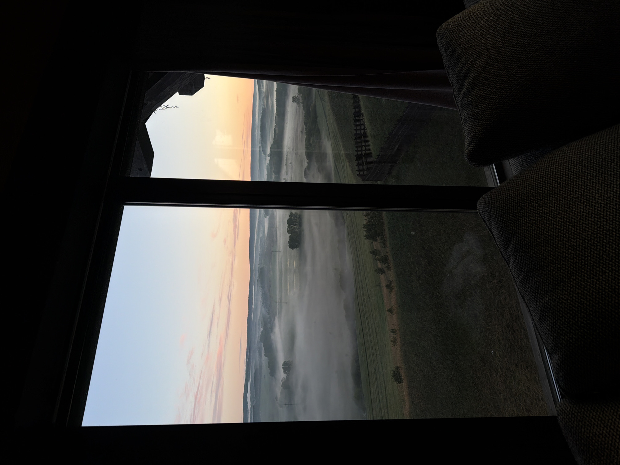 The image depicts a view through a window, likely from inside a room. The window is divided into two panes. Outside, a rural landscape is visible, shrouded in a thick layer of fog. The fog obscures much of the ground, but fields and rows of trees are partially visible through the mist. The sky is a mix of colors, with a pinkish-orange hue near the horizon, suggesting either sunrise or sunset. The window frame is dark, and a portion of a dark upholstered object, possibly a piece of furniture, is visible on the right side of the frame. The photo doesn't show any people, subjects, objects, actions, or interactions. The environment is a room and rural fogged view. The information does not suggest it is in Shamoni, Russia.