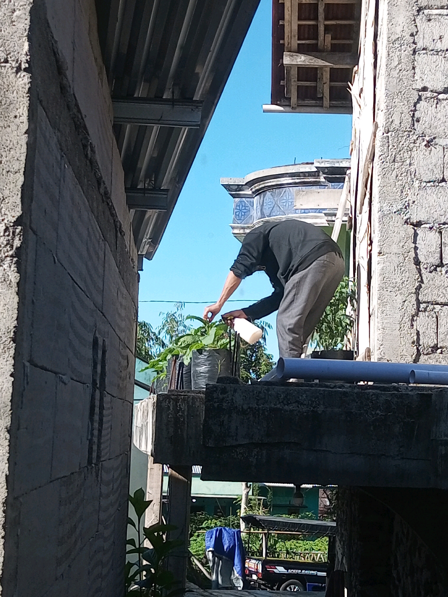 An individual, wearing a dark long-sleeved shirt and grey pants, is observed from a low-angle, side view, tending to a potted plant. The person is holding a white bottle, appearing to water or spray the plant. The plant is in a grey, cylindrical pot with a textured surface. The setting appears to be outdoors, with a clear blue sky visible in the background. Structures made of concrete blocks and corrugated metal roofing are present, suggesting an urban or semi-urban environment. A portion of a vehicle with "SPEED RACING" text is visible in the lower right, and blue tarp is also present. The overall scene conveys a sense of domestic or everyday activity within a developing or somewhat rustic area.