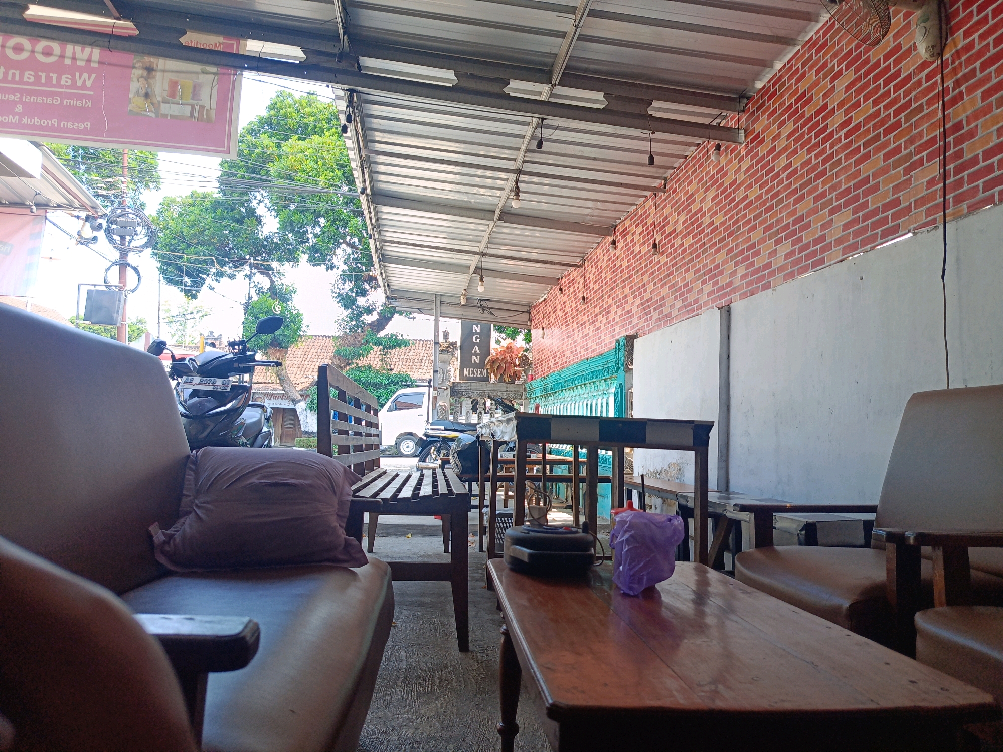 The image depicts an outdoor seating area, possibly a cafe or restaurant, in Candirejo, Indonesia. In the foreground, a brown leather armchair is visible on the left, with a brown throw pillow placed on its seat. To the right, a dark wooden coffee table occupies the center of the frame. On the table, a dark electronic device, a purple plastic bag, and a small red object are present. Behind the table, a wooden bench with dark planks is set up. Further back, a motorcycle is parked. The seating area is covered by a corrugated metal roof supported by a metal frame. A brick wall, constructed with red and orange bricks, forms the background on the right side of the image, with a concrete wall section below it. Trees with lush green foliage are visible in the background on the left. The ground appears to be paved.