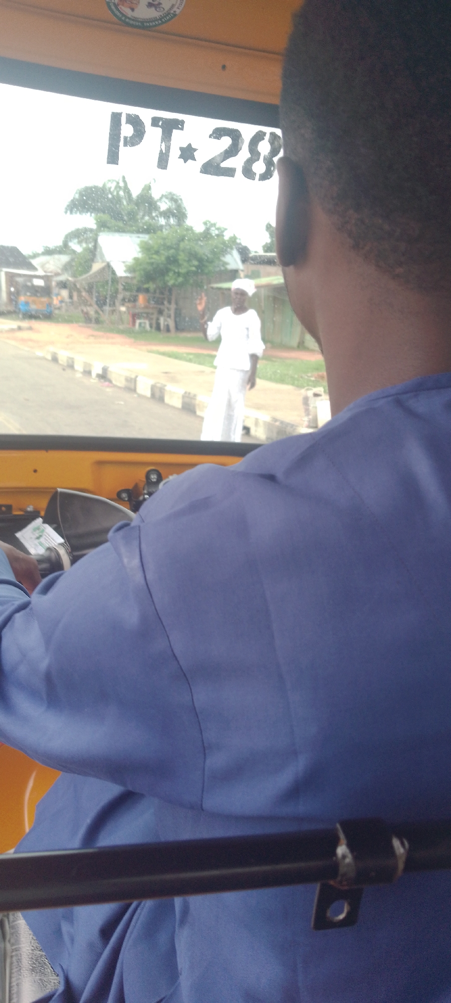The image shows the interior of a three-wheeled vehicle, likely a Keke Napep, in Jalingo, Nigeria. The perspective is from the driver's seat, looking forward through the windshield. A person's head and shoulder, wearing a blue garment, are visible in the foreground, obscuring a portion of the view.
Through the windshield, a woman dressed in white is standing on the side of a paved road, facing towards the vehicle and raising her right hand as if to signal. She is wearing a headscarf. Behind her, a residential area is visible with low-rise buildings, some with corrugated iron roofs, and scattered trees. A yellow three-wheeled vehicle is parked in the distance to the left.
Black stenciled lettering on the upper part of the windshield reads "PT-28". The road has a curb with black and white alternating markings. The overall scene depicts a street view in Jalingo.