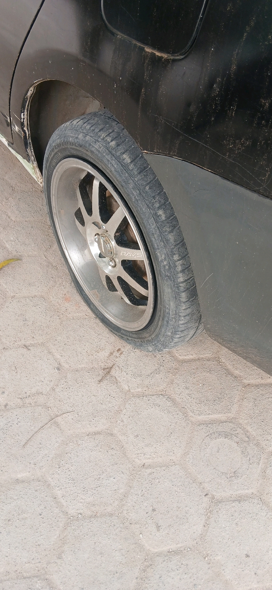 The image shows a close-up view of the rear passenger-side wheel and fender of a black car. The car's body is dirty and scratched, with some visible damage around the wheel well. The wheel itself is a silver alloy rim with black accents, and the tire is worn. The car is parked on a light-colored paved surface, possibly a street or parking lot, with a hexagonal pattern. The scene appears to be outdoors during the daytime, with neutral lighting. The image was taken in Metro, Indonesia.