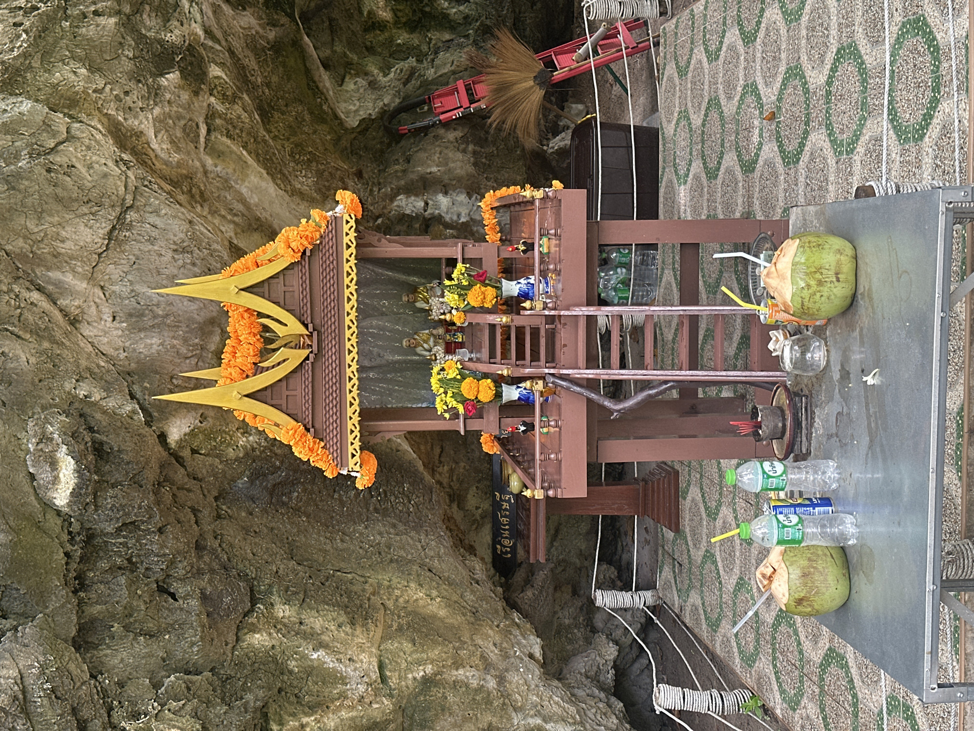 An outdoor shrine is set up within a cave in Ao Nang, Thailand. The shrine is made of dark wood and features a tiered roof decorated with orange marigold garlands and golden finials. Two statues are visible inside the shrine, along with floral offerings. A wooden ladder leads up to the shrine's platform. To the right of the shrine, a red stepladder leans against the cave wall, with a broom placed on it. The ground in front of the shrine is paved with hexagonal tiles. A metal table in the foreground holds two whole coconuts, one with a straw, and several plastic water bottles. Incense sticks are burning in a small holder on the shrine's lower level. The background consists of the rough, textured surface of the cave wall.
