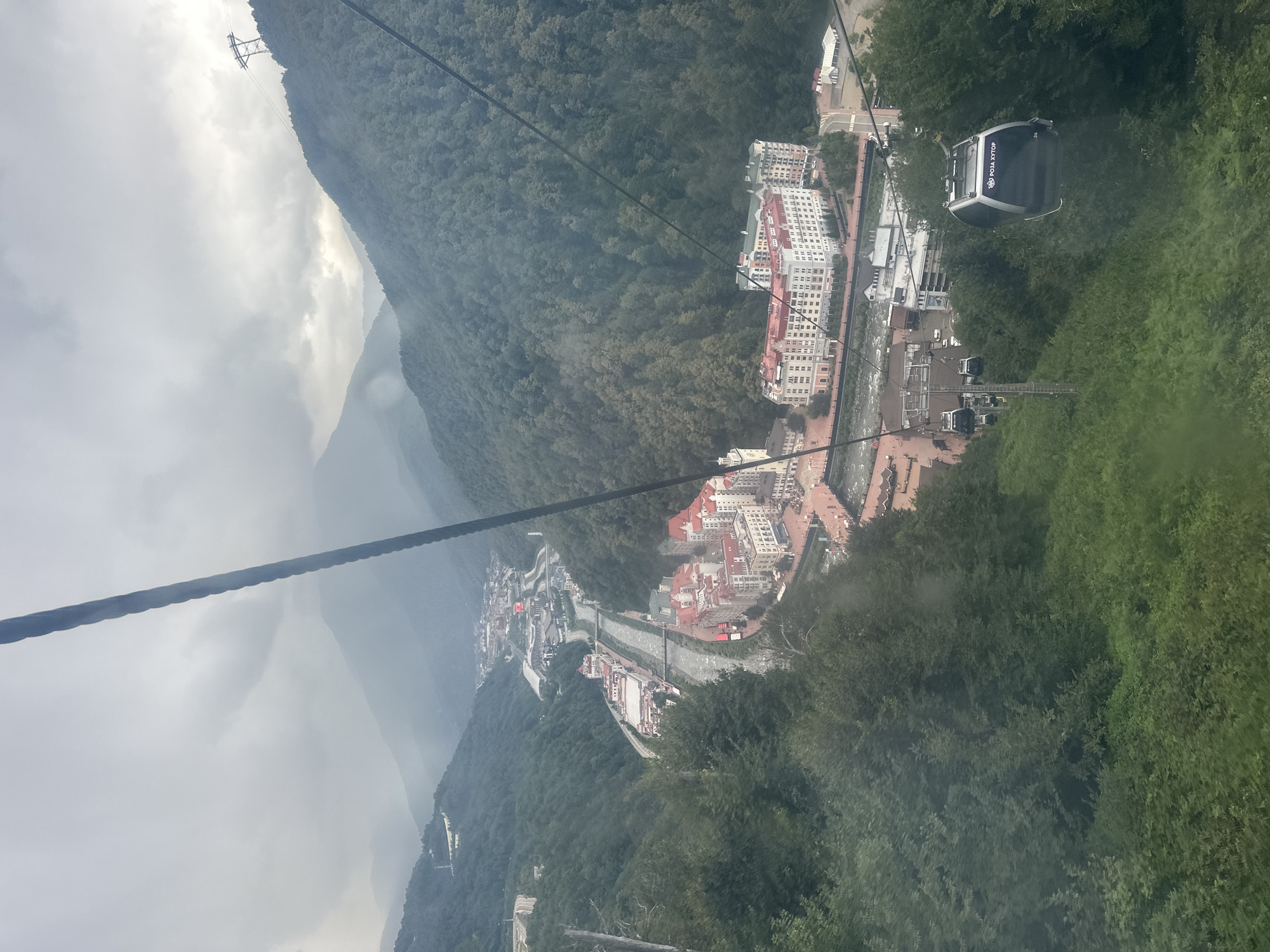 The image depicts a view from a cable car in Estosadok, Russia. The cable car is traveling down a mountain, offering a panoramic view of a valley below. The valley is filled with lush green trees, and a river flows through it. Several buildings are visible along the river, with red and white roofs. The sky is overcast, with clouds obscuring the mountain peaks in the distance. The cable car itself is modern and spacious, with large windows that allow for an unobstructed view of the surroundings. The scene suggests a popular tourist destination, likely a ski resort given the mountainous terrain and cable car infrastructure.