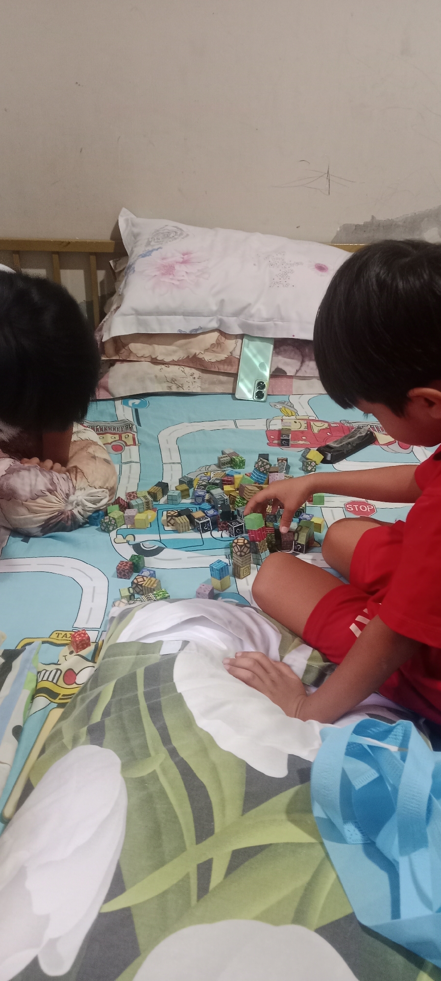 In Banyuraden, Indonesia, two children are playing on a bed. The bed is covered with a blue sheet depicting a city map with roads and buildings. Scattered across the sheet are numerous small, colorful blocks, which the children are arranging and building with. One child, wearing a red shirt and shorts, is actively engaged with the blocks, while the other child, partially obscured, appears to be also interacting with the toys. Pillows are stacked at the head of the bed, and a light-colored wall serves as the backdrop. A mobile phone is placed on the bedsheets near the pillows. The overall environment suggests a casual, indoor playtime activity.