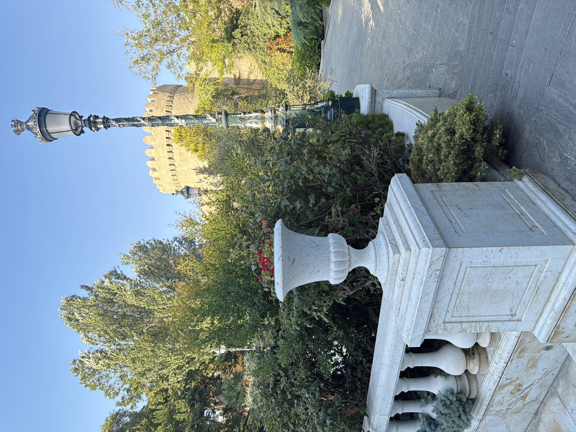 This image captures a view of the Maiden Tower in Baku, Azerbaijan, with lush greenery and architectural elements in the foreground. In the foreground, a white marble balustrade with ornate balusters lines a pathway. A large white marble urn filled with red flowers sits on a pedestal to the left of the balustrade. To the right of the urn, a dark green, ornate lamppost stands tall, its intricate details illuminated by sunlight. Behind the lamppost, a cobblestone pathway curves away, leading towards the Maiden Tower. The tower itself, a cylindrical stone structure with crenellated battlements, stands in the background, partially obscured by dense green foliage. The sky above is a clear, vibrant blue, indicative of a sunny day. The scene suggests a well-maintained park or garden area within the historic precinct of Baku.