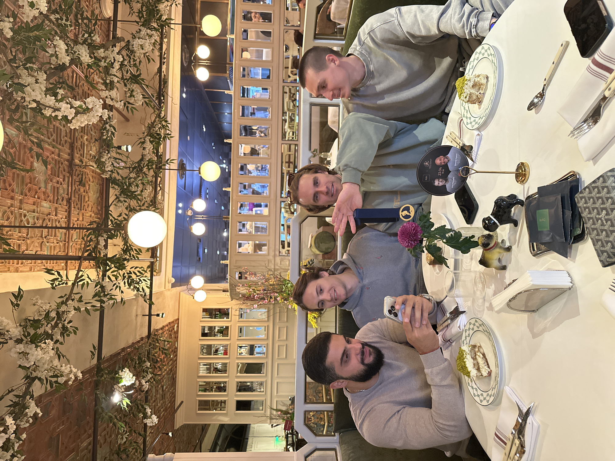 Four young men are seated at a round table in a restaurant in Moscow, Russia. They are likely at a celebratory event. On the table are plates with slices of pistachio cake, napkins, silverware, a phone, and decorative objects, including flowers. The restaurant has a mirrored wall.