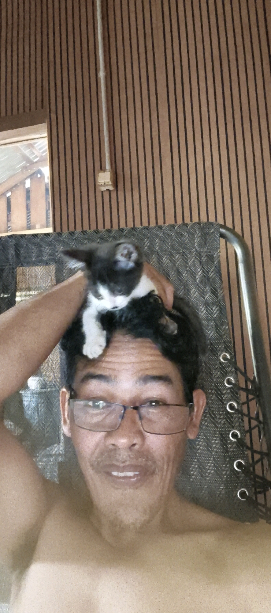 The image shows a close-up, selfie-style shot of a person with a kitten on their head. The person, who appears to be an adult male, is wearing glasses and has a slight smile. The kitten is black and white and is positioned on top of the person's dark hair. The background consists of vertical wooden planks. The scene appears to be indoors, possibly in a home. The lighting is moderate, and the image has a slightly grainy quality. The provided hints do not offer any information about Muaro Jambi, Indonesia.