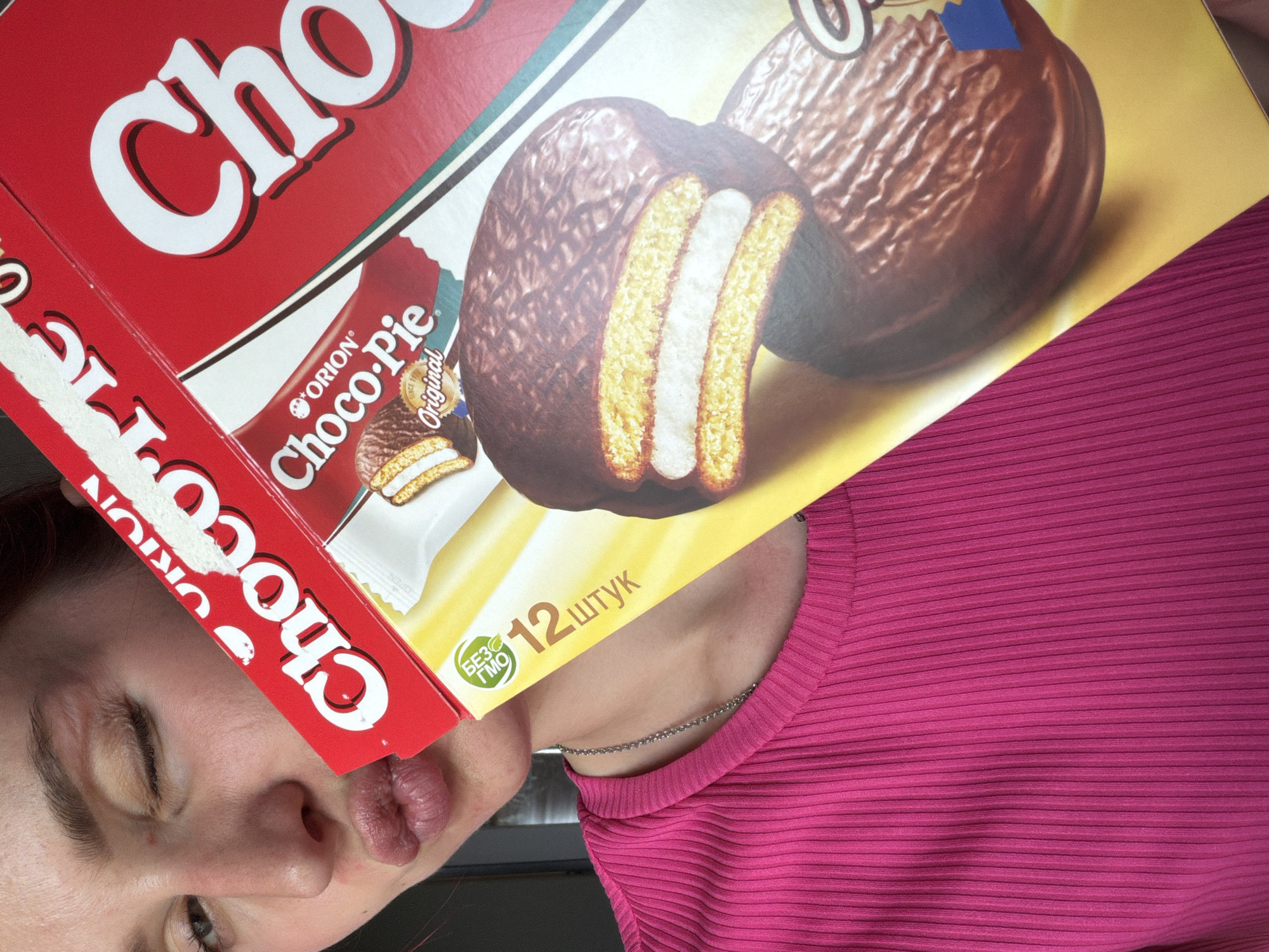A person with fair skin, wearing a bright pink, horizontally ribbed top, is partially visible. Their face is angled downwards to the left, with one eye closed and the other partially open. Their lips are puckered, suggesting a playful expression. They appear to be holding a box of "Orion Choco-Pie Original" in front of their face, partially obscuring it. The box is red, yellow, and white, with images of the chocolate pies and text in Cyrillic script. The image is brightly lit and slightly angled. There is no visible information in the image about the city of Chaikovsky, Russia.