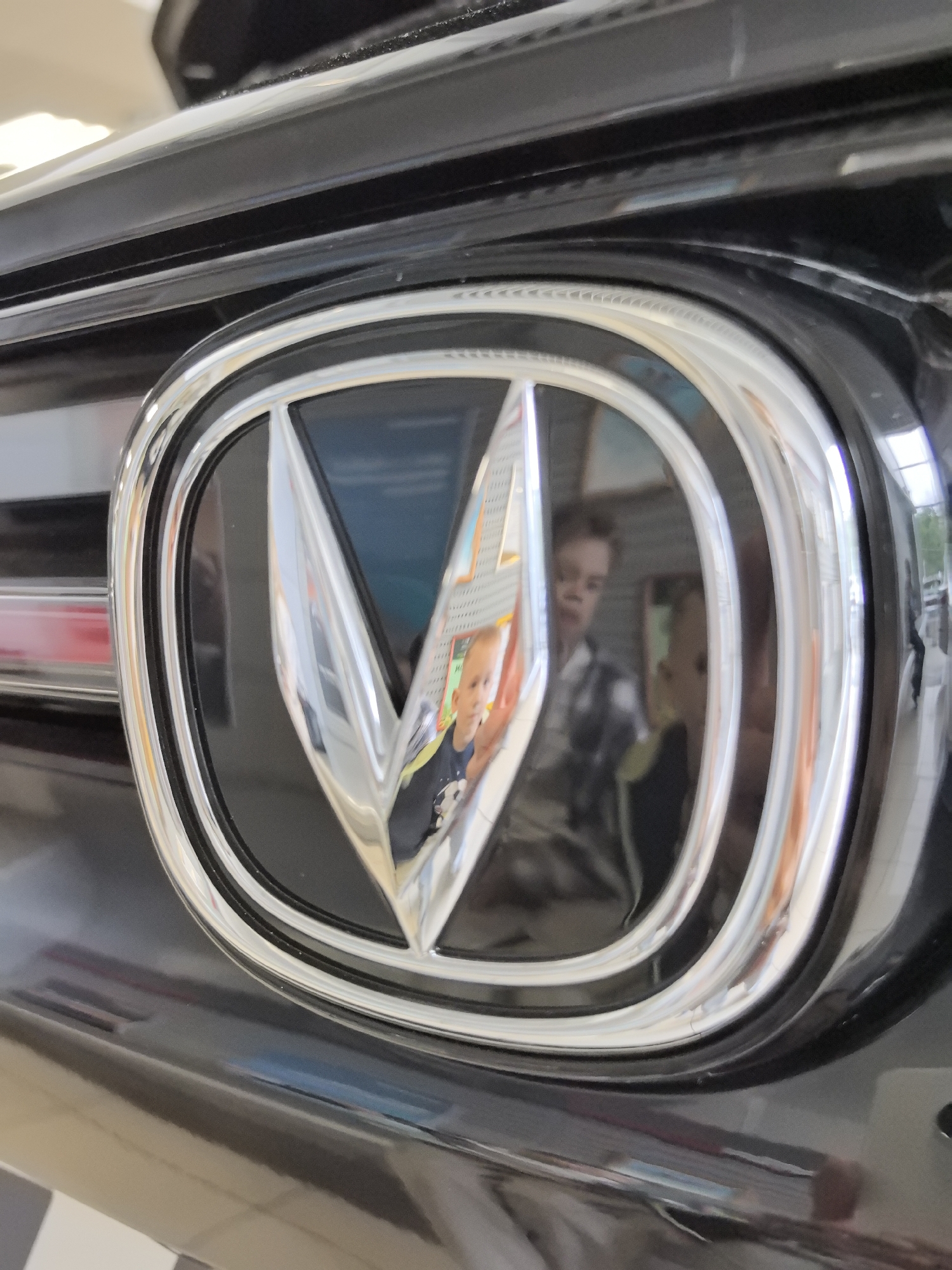The image shows a close-up of a car's emblem, which appears to be a stylized "V" shape within a silver oval frame, set against a black car body. The emblem is reflecting two children, a younger child in the foreground wearing a dark shirt with a graphic, and an older child or young man in the background wearing a plaid shirt. The reflection also shows some display posters or signs behind the children. The background outside the reflection suggests an indoor environment, possibly a showroom or a garage, with a floor that has a checkered pattern. The specific location in the city of Perm, Russia, cannot be determined from the visual information in the image.