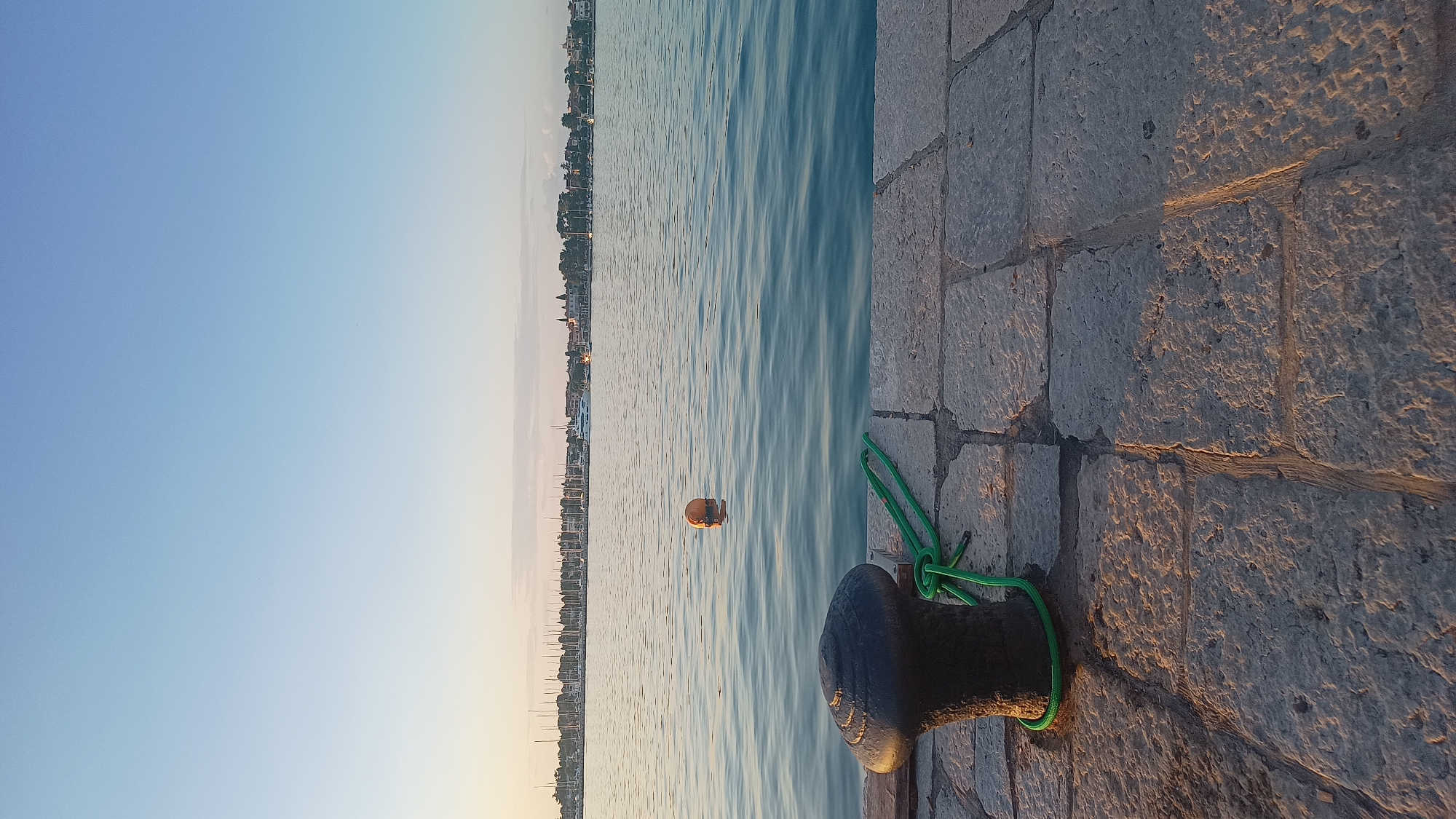 The image depicts a static waterscape scene at a marina in what is identified as Umag, Croatia, likely during either sunrise or sunset due to the warm, low-angle light. The sky occupies the top two-thirds of the vertical frame, exhibiting a clear gradient from a pale, light blue at the zenith to a warmer, more yellowish-orange hue near the horizon, indicative of an atmospheric transition.
The water surface below the sky is calm with minor ripples, reflecting the ambient light and appearing in shades of blue and muted green. In the midground, an orange spherical buoy is visible, partially submerged and casting a subtle reflection on the water.
The foreground features a stone-paved pier or dock extending from the lower edge of the frame. The paving consists of irregularly shaped, flat stone blocks with visible grout lines. On the left side of the foreground, a dark, heavy-duty metal bollard is anchored to the paving. A bright green, braided rope is coiled around the base of the bollard and extends onto the stone surface to its right. The low-angle sunlight illuminates the textured surface of the stones and the bollard, creating defined shadows.
Across the water in the background, a distant coastline is visible. This coastline is densely populated with buildings, trees, and a marina filled with numerous docked sailboats, identifiable by their prominent masts forming a dense linear pattern. A larger white yacht is also discernible among the smaller vessels. No individuals are present in the frame.