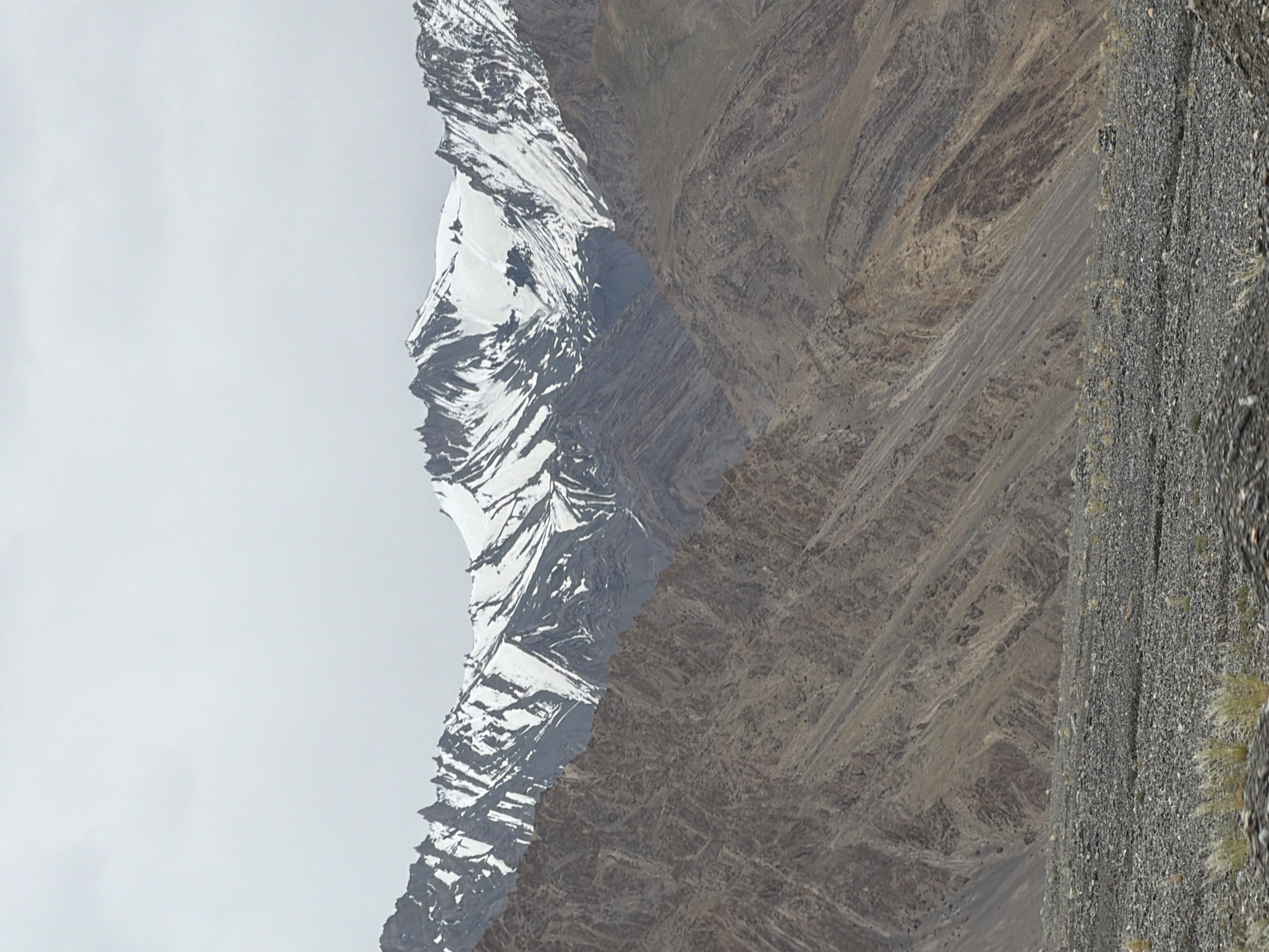 A wide shot captures a rugged, mountainous landscape with a rocky, desolate foreground. In the distance, towering peaks are capped with snow, their jagged ridges contrasting with the muted, cloudy sky. The middle ground features steep, rocky slopes exhibiting a layered geological structure, with shades of brown and grey dominating the terrain. Patches of sparse, dry vegetation are scattered across the rocky foreground, adding a touch of muted color to the otherwise barren scene. The overall impression is one of an isolated and harsh natural environment.