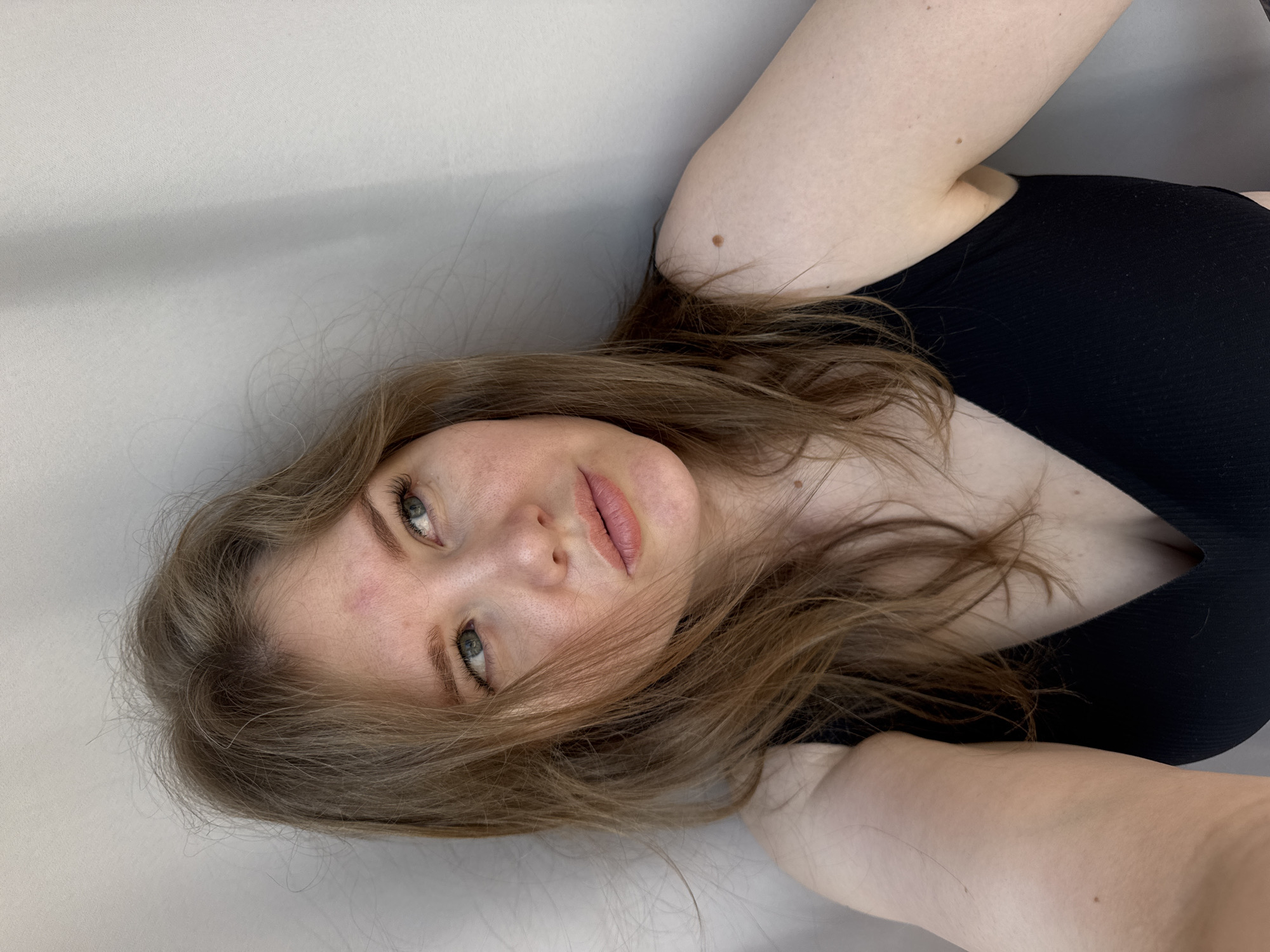 The image shows a woman lying down. She has fair skin, brown hair, and green eyes. She is wearing a black top with a low neckline. Her arms are visible, and there is a mole on one of them. The background is a plain, light-colored wall. There is no indication of a specific location in Gornyi, Russia, or any other identifiable geographical feature.
