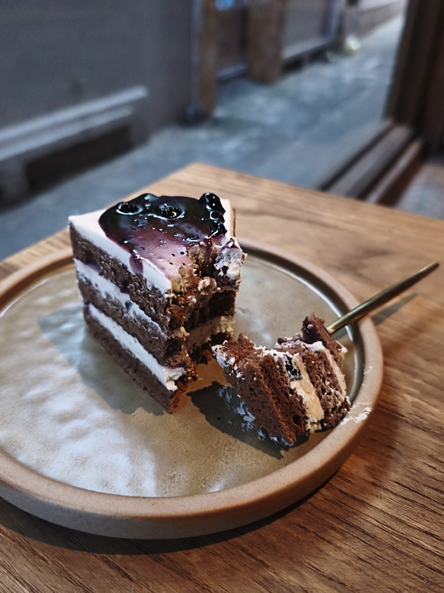 A slice of chocolate cake with white frosting and blueberry topping sits on a round ceramic plate on a wooden table. A fork with a gold handle is inserted into the cake, with a small piece of cake on its tines. The background is blurred, suggesting an indoor setting possibly a cafe or restaurant. The location is identified as Bac Nha Trang Ward, Vietnam, based on the provided information.