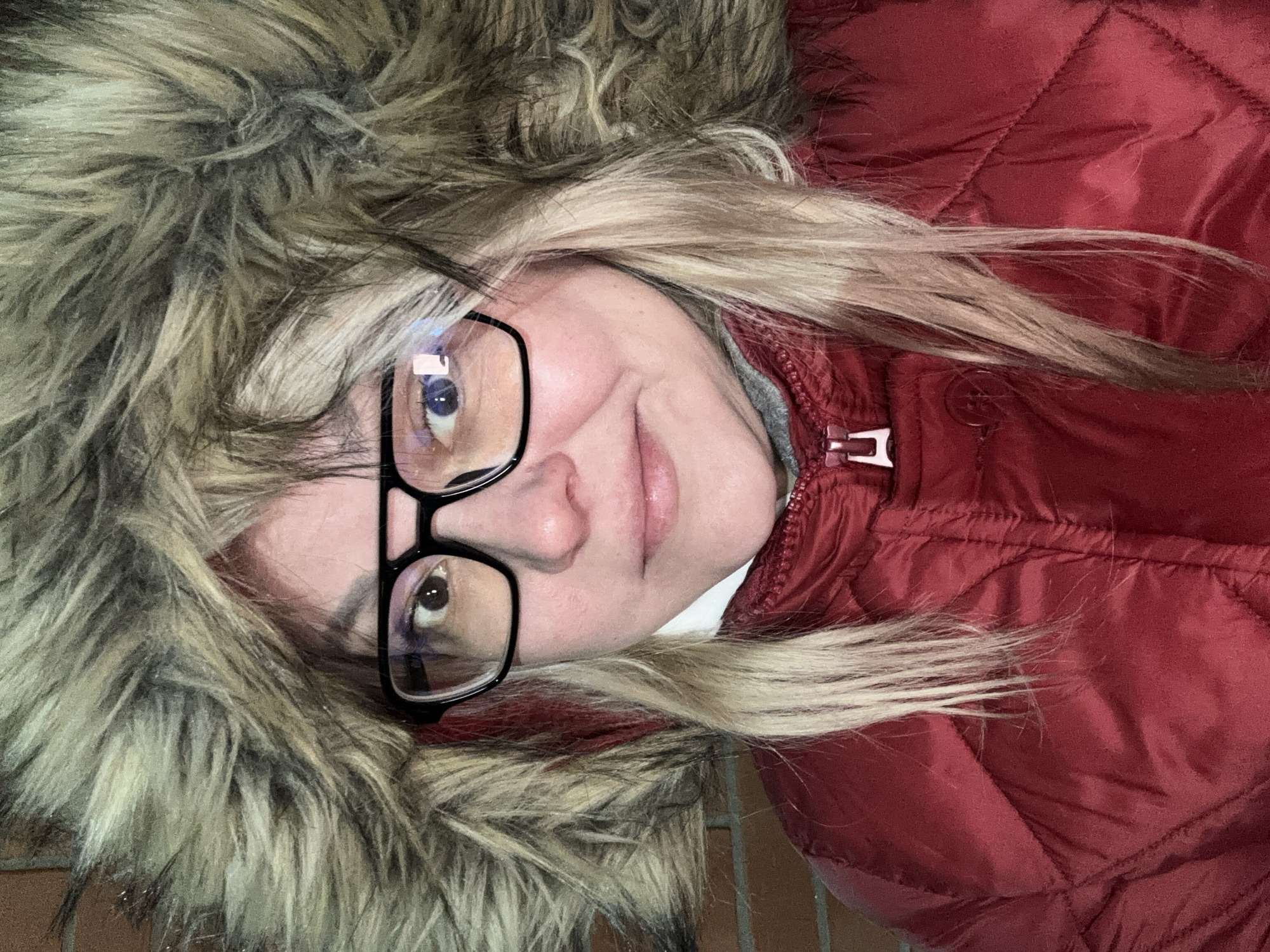 A close-up photograph taken in Русский Вожой, Russia, depicts an adult female subject looking directly at the camera. The individual is wearing a red quilted winter jacket featuring a prominent, multi-toned faux fur hood in shades of brown and grey. Beneath the red jacket, layers of grey and white fabric are visible around the neck area. The subject has light skin, brown eyes, and wears black-framed eyeglasses, which show reflections from ambient light sources. Her hair, which is predominantly blonde with some darker and red streaks, frames her face and emerges from under the hood. Her lips are closed with a slight upward curve, indicative of a subtle smile. The background is an out-of-focus reddish-brown brick wall.