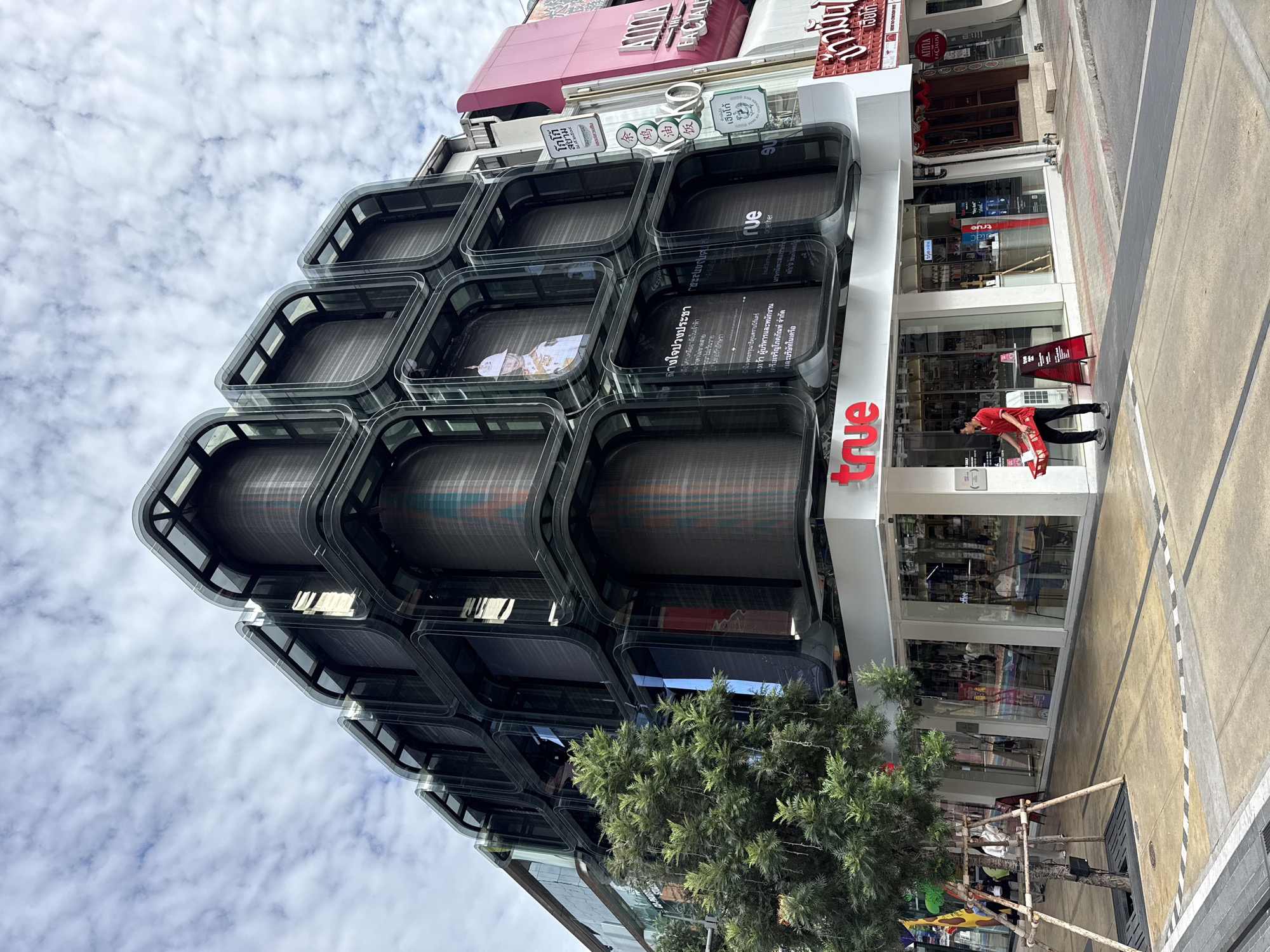 This is a street-level, eye-level shot of a modern, multi-story building with a unique, cubed facade covered in digital screens. The building is in Bangkok, Thailand. The facade is a dark, translucent material with rounded edges forming a grid of large, rectangular panels. Many of these panels display digital content, including images of a Thai royal figure, text in Thai, and abstract patterns. The ground floor features a retail space with large glass windows, branded "true" with a red logo. Inside, displays and seating are visible. A man in a red shirt and black pants is walking on the sidewalk, carrying a red basket. The street is paved with concrete and asphalt, and there are sidewalks with some yellow and white painted markings. A green tree with a visible wooden support structure is planted on the sidewalk. Other buildings with signage in Thai and English are visible to the right of the main building. The sky is partly cloudy.