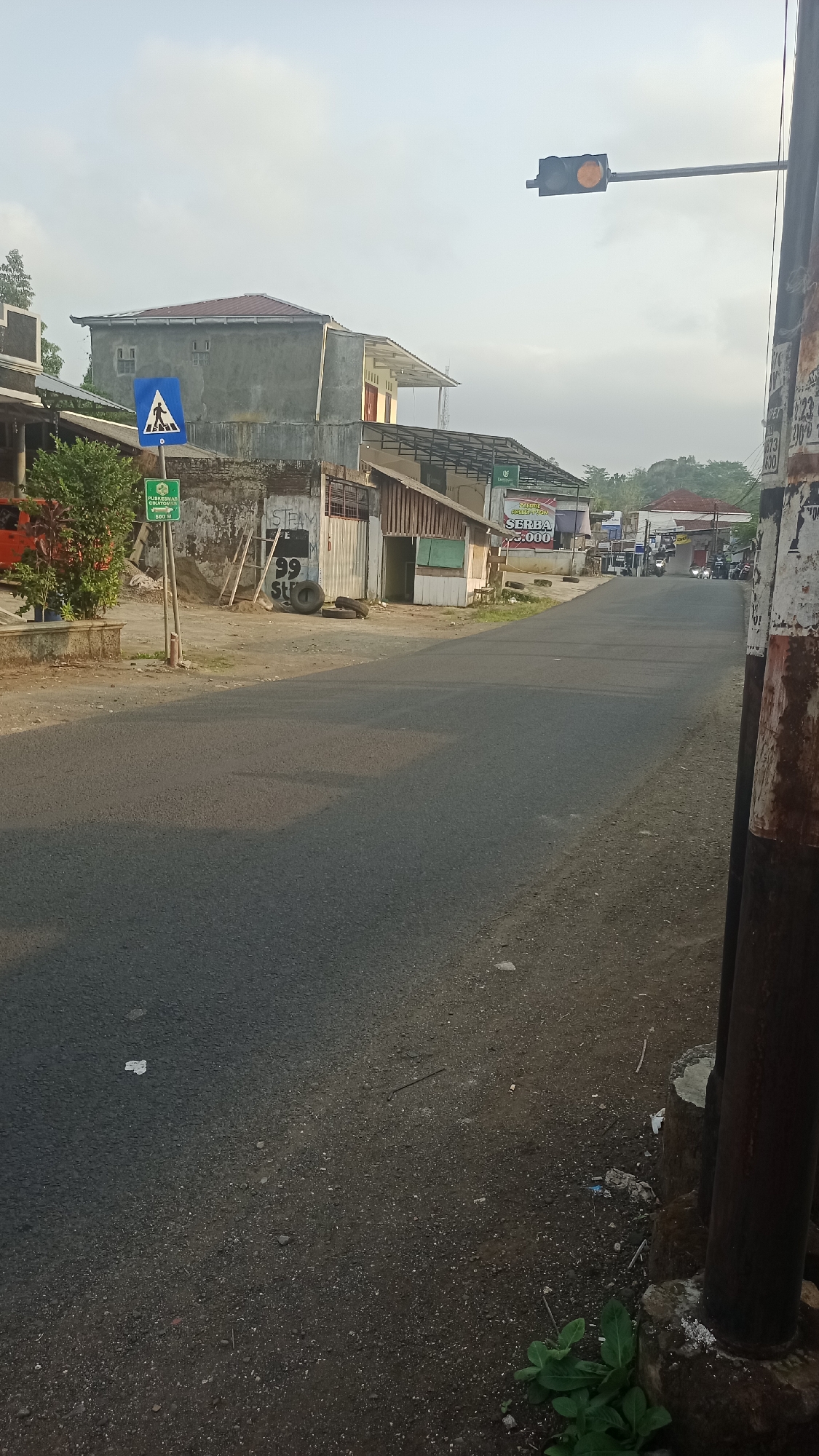The image depicts a street scene in Karyamandala, Indonesia, likely during the late afternoon or early morning due to the soft lighting. On the left side of the road, a pedestrian crossing sign is visible, along with a green sign indicating "PUSKESMAS CIHATEUL" with an arrow and "500 M". Adjacent to the sign is a dense bush with red-tinged leaves. Behind this, a building with a gray stucco exterior and a reddish-brown tiled roof is situated. Further down the street, several small buildings with corrugated metal roofs and wooden walls line the road, some appearing to be shops or stalls. One building has a sign that reads "SERBA 5.000". A traffic light with an illuminated orange signal hangs over the intersection ahead. The road itself is paved asphalt, with some debris scattered on the shoulder. In the distance, the road curves and several motorbikes and vehicles are visible, suggesting ongoing traffic. A large, weathered metal pole stands on the right side of the road, partially covered in peeling paint and what appears to be a poster or graffiti. The sky is overcast with a pale, diffused light.