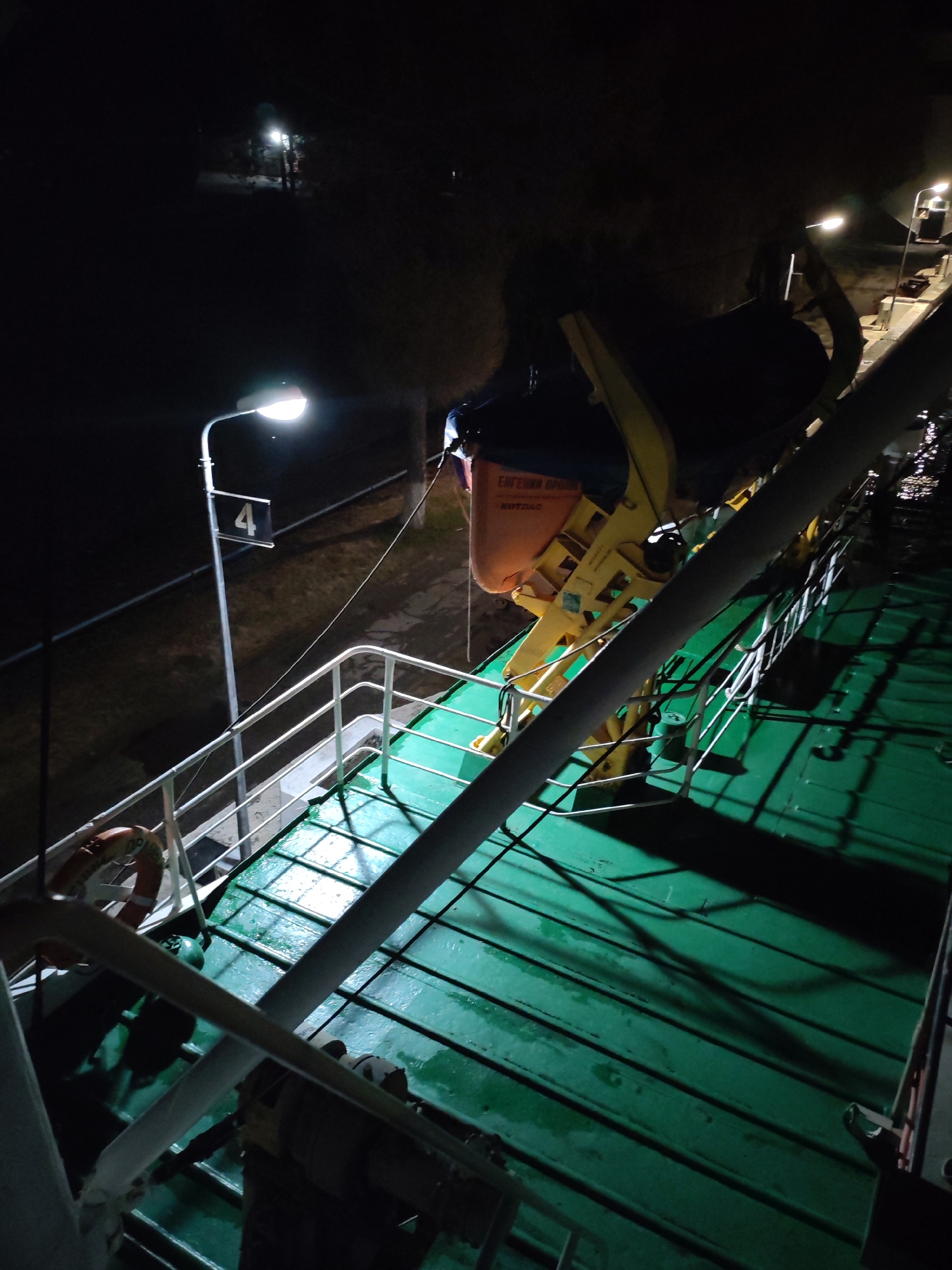 The image depicts a view from the deck of a vessel at night. The deck is green and appears wet. A davit arm for launching a life raft is visible, with an orange life raft container attached. A white railing runs along the side of the deck. A white light post is visible on the shore, marked with the number "4". A life ring is attached to the railing. In the background, there are dark trees and other light posts.