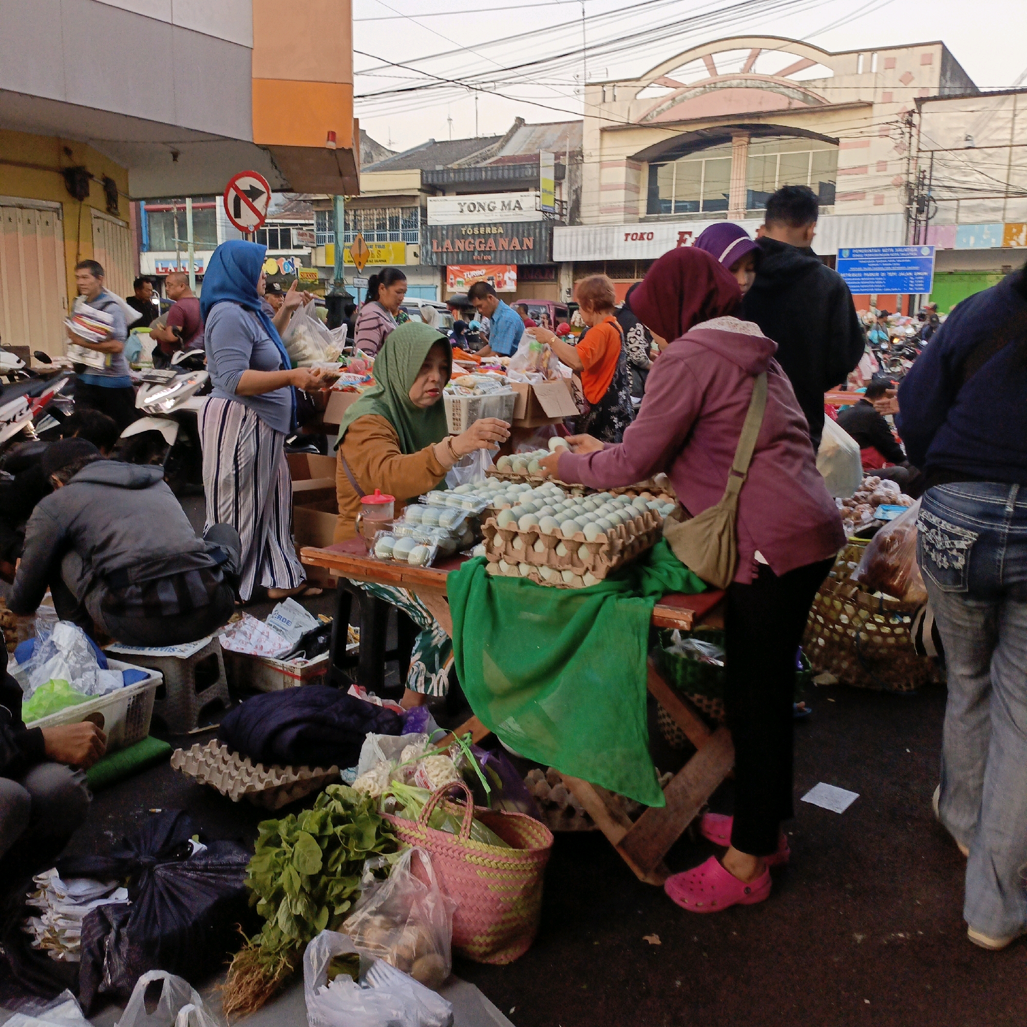 The image captures a bustling outdoor market scene in Salatiga, Indonesia. The foreground is dominated by a makeshift stall with wooden planks supporting various goods. Piles of green vegetables, including leafy greens with roots, are prominent on the left. Several plastic bags contain what appear to be potatoes or similar produce. A pink and green woven basket sits nearby, holding more of the green vegetables. Egg cartons, filled with pale blue eggs, are displayed on the stall.
Several individuals are interacting within the market. A woman in a green headscarf and brown top is examining a customer's hand near the egg display. Another woman, wearing a purple headscarf and a pink jacket, is standing behind the stall, her face obscured. A person in black pants and a pink clog is visible on the right side of the stall. Further in the background, people are seen browsing and purchasing items. Motorcycles are parked behind the stalls, indicating transportation used by shoppers and vendors. The background reveals buildings with signage, including "YONG MA" and "TOKO LANGGANAN," suggesting a commercial area. The sky is overcast, and the overall lighting suggests it is either early morning or late afternoon. The ground is a dark, rough surface, likely asphalt or dirt, typical of a street market.
