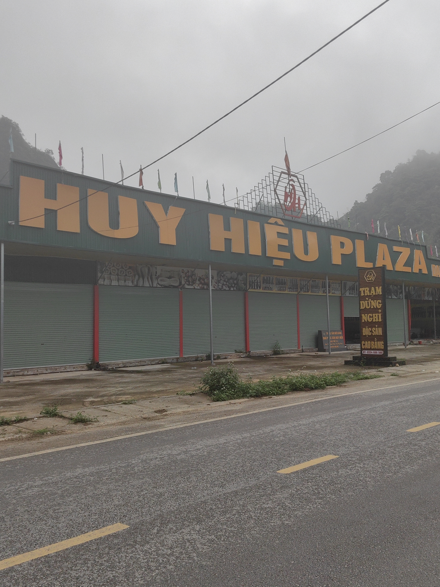 The image depicts the "Huy Hiệu Plaza" building, possibly a roadside service stop or a collection of shops, located on a wet asphalt road with yellow lane markings in Quảng Uyên, Vietnam. The building features a large green facade with prominent yellow lettering that reads "HUY HIỆU PLAZA." Additional, smaller signage in front of the building promotes the "Tram Dừng Nghi Đặc Sản Cao Bằng," which appears to advertise local Cao Bằng specialties, along with a phone number and information about Dao PC Sen Huy Hiệu. The building itself has multiple storefronts behind closed, green roller shutters with red vertical stripes. Flags are visible atop the roof, with a decorative structure incorporating the letters "DA" above the name. A mountainous, hazy background suggests the location is situated in or near a mountainous region. There are some weeds and greenery growing on the pavement in front of the building. The weather appears overcast and damp.