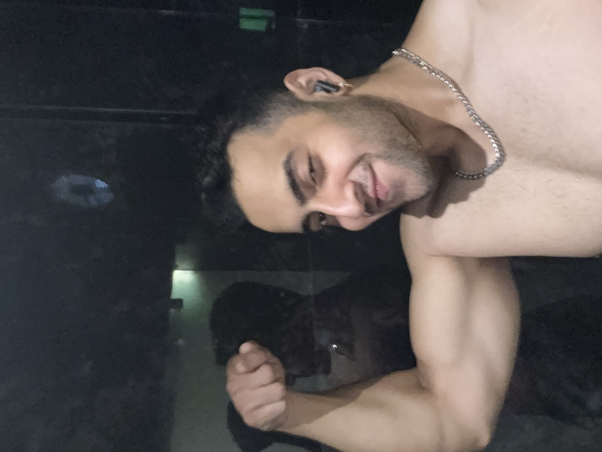 A shirtless man with dark hair and a silver chain around his neck is flexing his bicep. He is wearing an earbud in his left ear. The background is dark with some light reflections. There is no information in the image to indicate that it was taken in Kolkata, India.