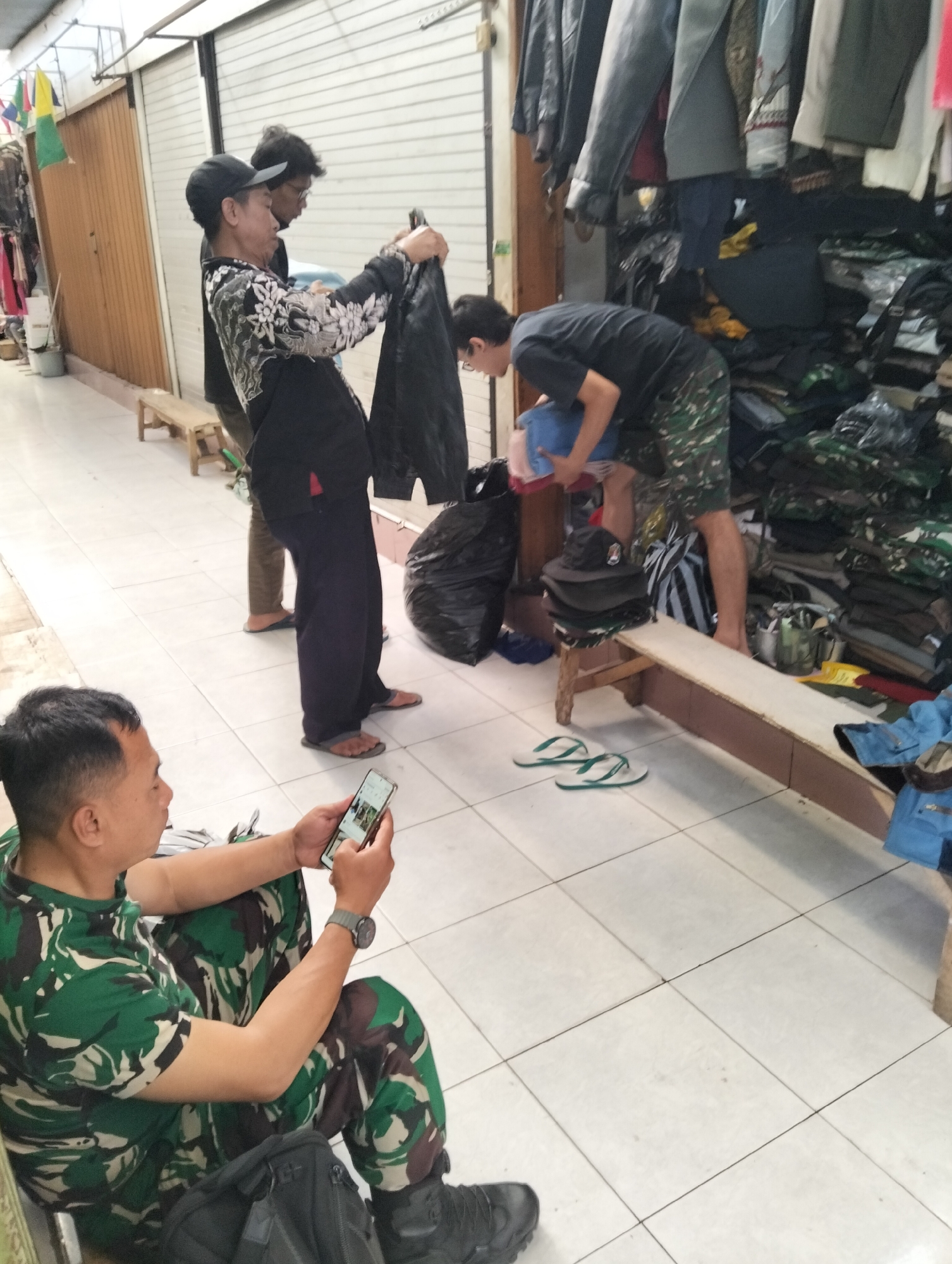 In Magelang, Indonesia, a group of men are seen in what appears to be an outdoor market or shop. One man in the foreground, dressed in a green camouflage uniform, is seated and looking at his smartphone, holding it in his left hand. He is wearing a wristwatch on his left wrist. Beside him on the floor is a black backpack.
In the background, three other men are present. One man, wearing a black baseball cap and a patterned shirt, is holding a dark plastic bag. He is standing next to another man who is partially obscured. Further back, a third man, wearing a black t-shirt and camouflage shorts, is bending over and appears to be holding or sorting some blue and pink items.
The environment is tiled and appears to be an aisle with various items for sale, including clothing hanging from racks and stacked on shelves. A small wooden bench is visible. The overall scene suggests a casual and possibly commercial setting.
