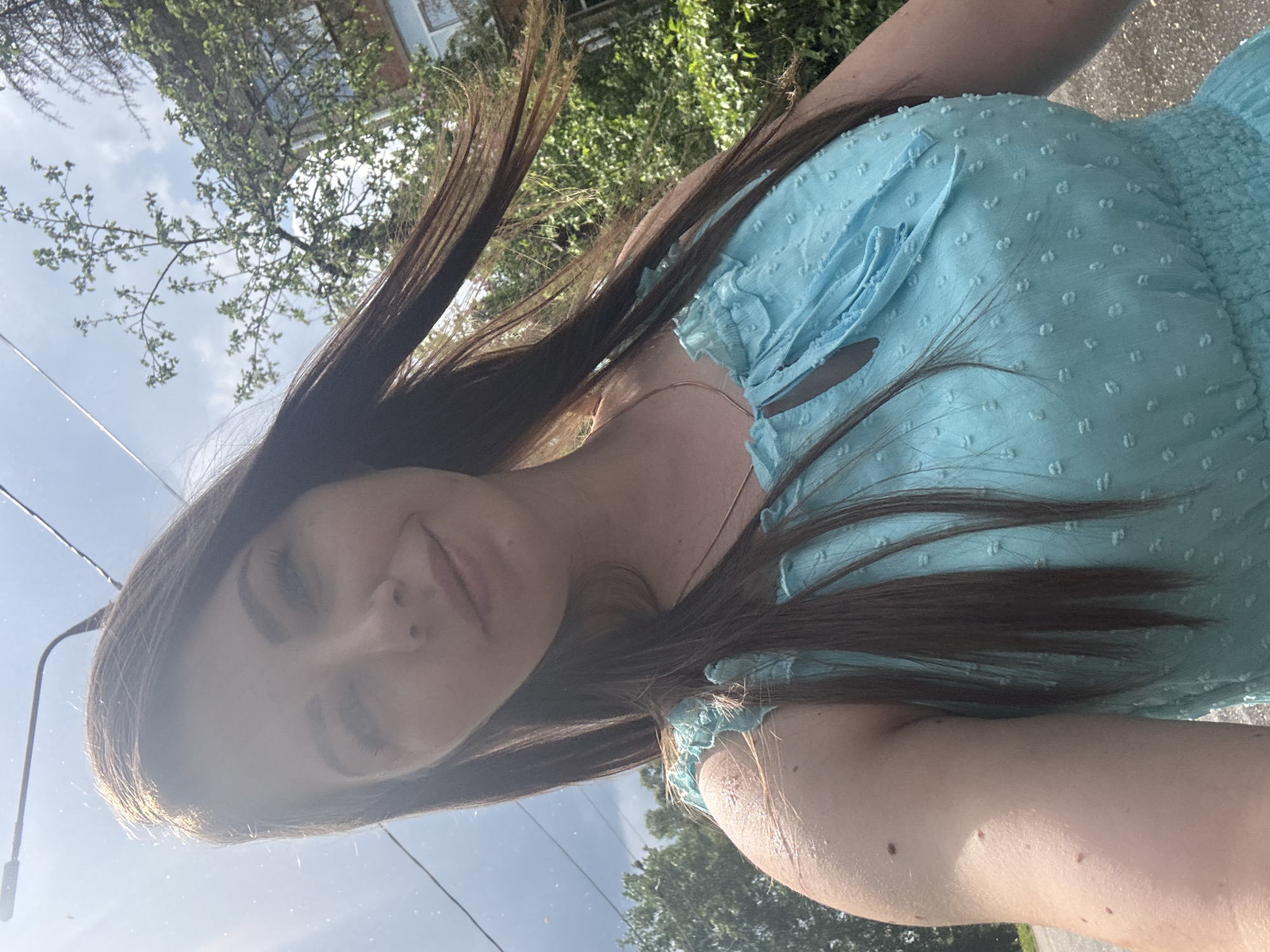 A selfie of a woman is taken outdoors, appearing to be on a street. The woman has long, dark hair and is wearing a light blue dress. In the background, there are trees and a building with a light-colored facade. The location cannot be determined to be Shevlyakovo, Russia from the image alone.
