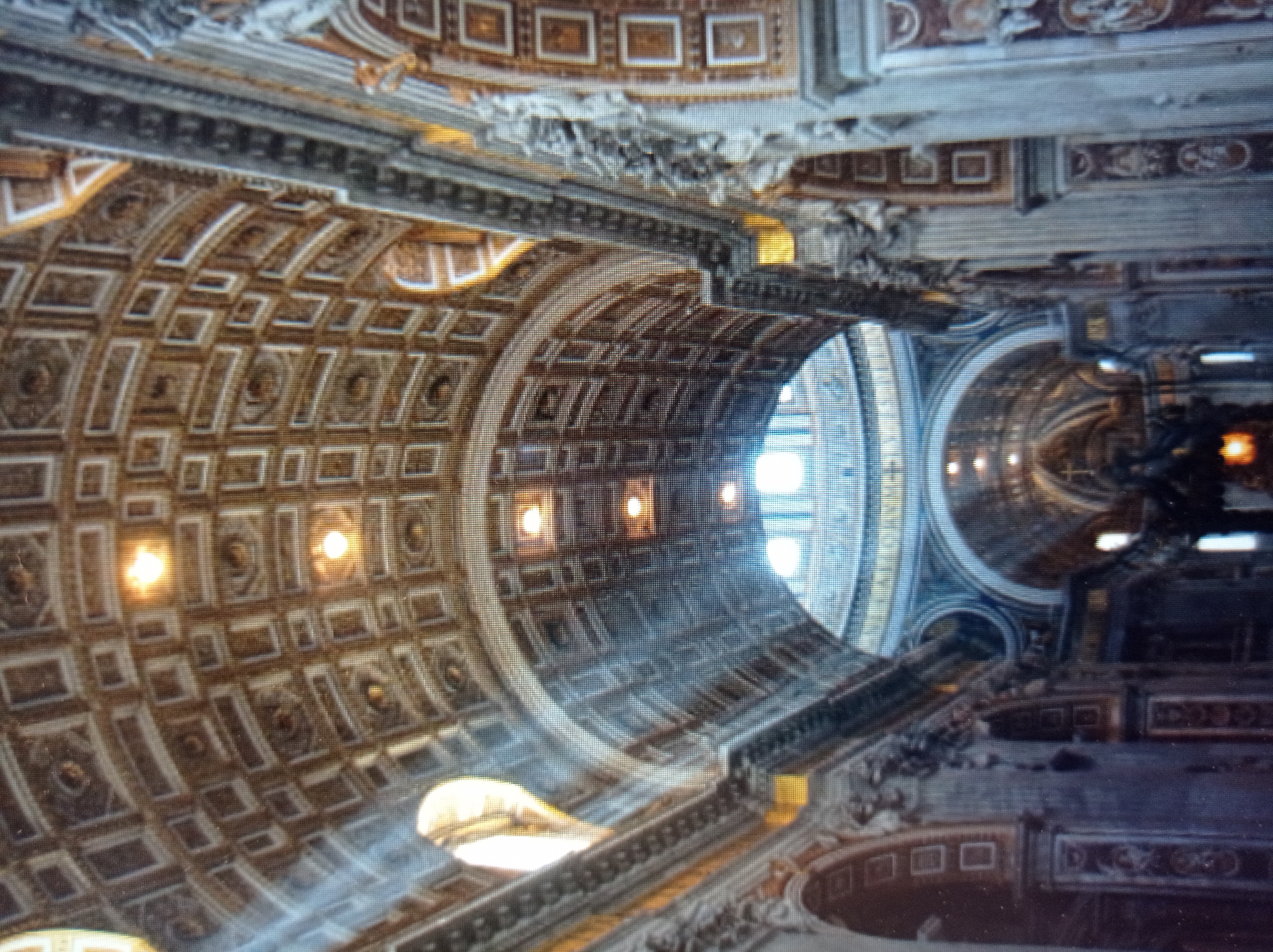 The image depicts the interior of a grand, ornate building, likely a cathedral or basilica, with a high, arched ceiling adorned with intricate geometric patterns and illuminated by multiple light sources. The architecture features ornate columns, decorative friezes, and arched doorways or windows. Sunlight streams in from large openings, casting light and shadow across the interior. The style suggests European classical or baroque architecture. There are no people visible in the image. The image quality is low resolution and pixelated. This scene is not located in Mukachevo, Ukraine; it is Saint Peter's Basilica in Vatican City.