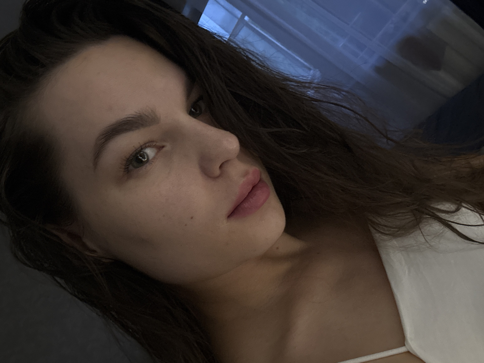 The image is a close-up, low-angle selfie of a young woman with long, dark brown hair. She is wearing a white top with thin straps. Her head is tilted, and she is looking off to the side with a slight gaze. Her skin appears to be fair with some subtle freckles. Her lips are slightly parted and have a soft pink hue. The background is dark and out of focus, with a hint of blue light coming from what appears to be a window with sheer curtains on the right side of the frame. The overall lighting is dim, casting shadows that accentuate the contours of her face. The location is Chaikovsky, Russia.