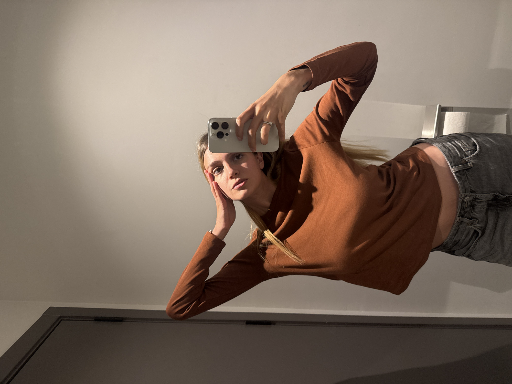 A young woman is taking a selfie in what appears to be a bathroom. She is holding a smartphone with three rear cameras in her right hand, positioned slightly above her forehead, and resting her left hand against her left cheek. She is wearing a long-sleeved brown top and faded gray jeans. Her blonde hair is partially pulled back. The background consists of a white wall, a partially visible silver fixture above the toilet (likely a toilet paper holder), and the upper portion of a dark gray door frame along the bottom of the frame. The scene suggests an indoor setting, possibly a residential bathroom. Without further context, no location in Tbilisi, Georgia can be confirmed.