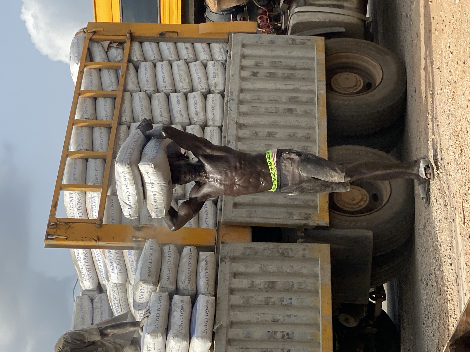 An unclothed man stands on a truck loaded with "MANGAL CEMENT" bags, in Adankolo, Nigeria. He balances a bag of cement on his head with both hands. The truck is yellow and grey and has two visible tires. A yellow ladder leans against the cement bags on the left side of the truck. The environment appears to be outdoors.
