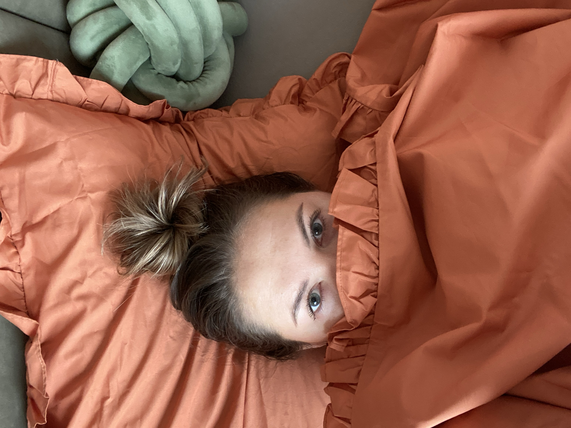 A woman is lying on a bed, with an orange ruffled sheet partially covering her face. Her eyes are visible above the sheet. Her hair is pulled back into a messy bun. A knotted, green pillow is visible in the upper left corner of the frame, resting on a grey surface. The setting appears to be indoors. The location, Izhevsk, Russia, cannot be confirmed from the given image.