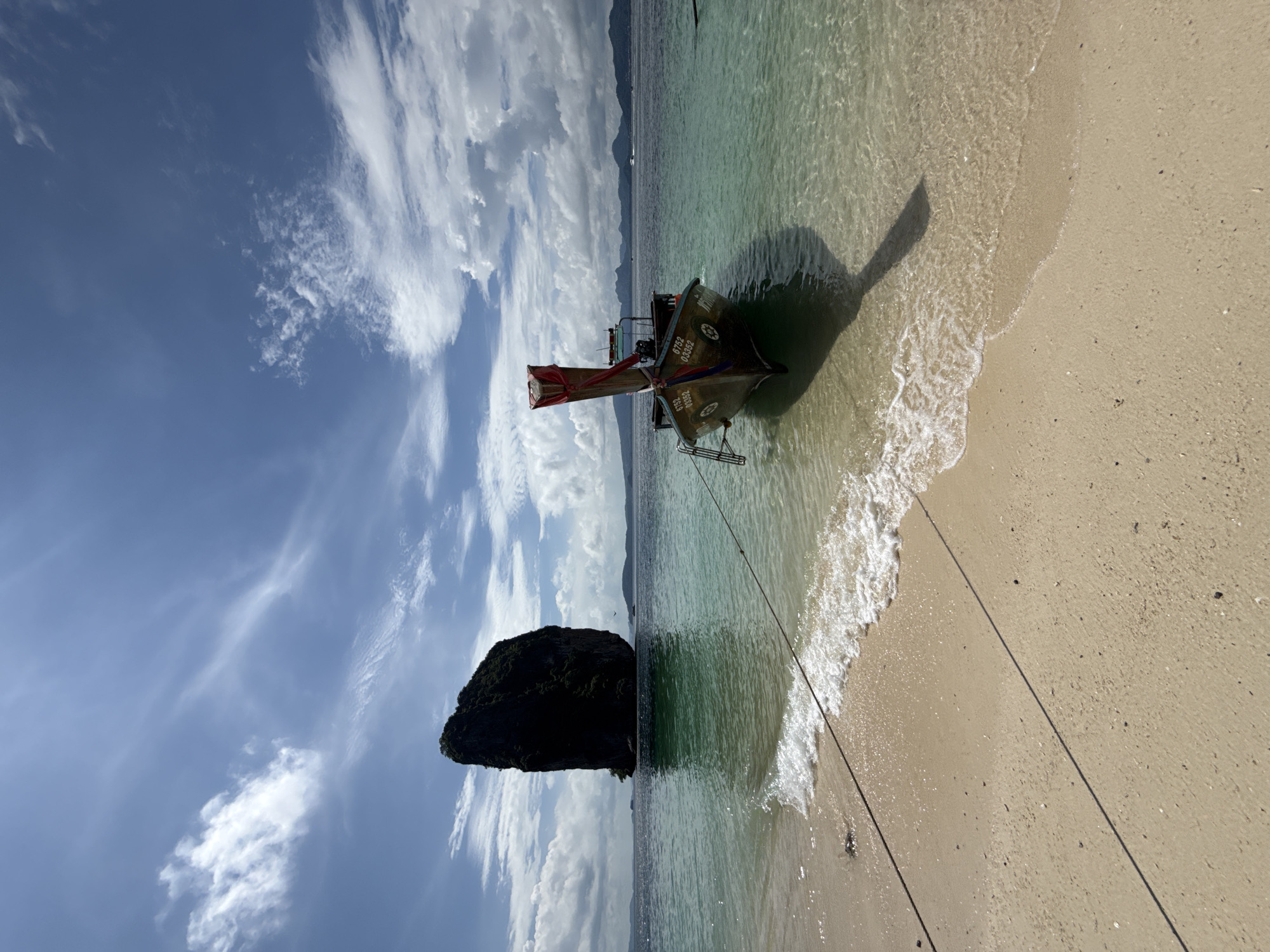 This is a medium-wide shot of a tropical beach with a clear, turquoise ocean and a prominent limestone karst formation rising from the water in the distance. The foreground features a sandy beach with gentle waves lapping at the shore. A traditional longtail boat is anchored in the shallow water, its hull adorned with numbers. The boat is tied to the shore with a rope that stretches across the sand and into the water. In the background, the karst island is silhouetted against a bright blue sky dotted with wispy white clouds. The overall scene depicts a serene and picturesque coastal environment, likely in or near Ban Ao Nang, Thailand, a region known for its dramatic limestone cliffs and clear waters. There are no people visible in the image.