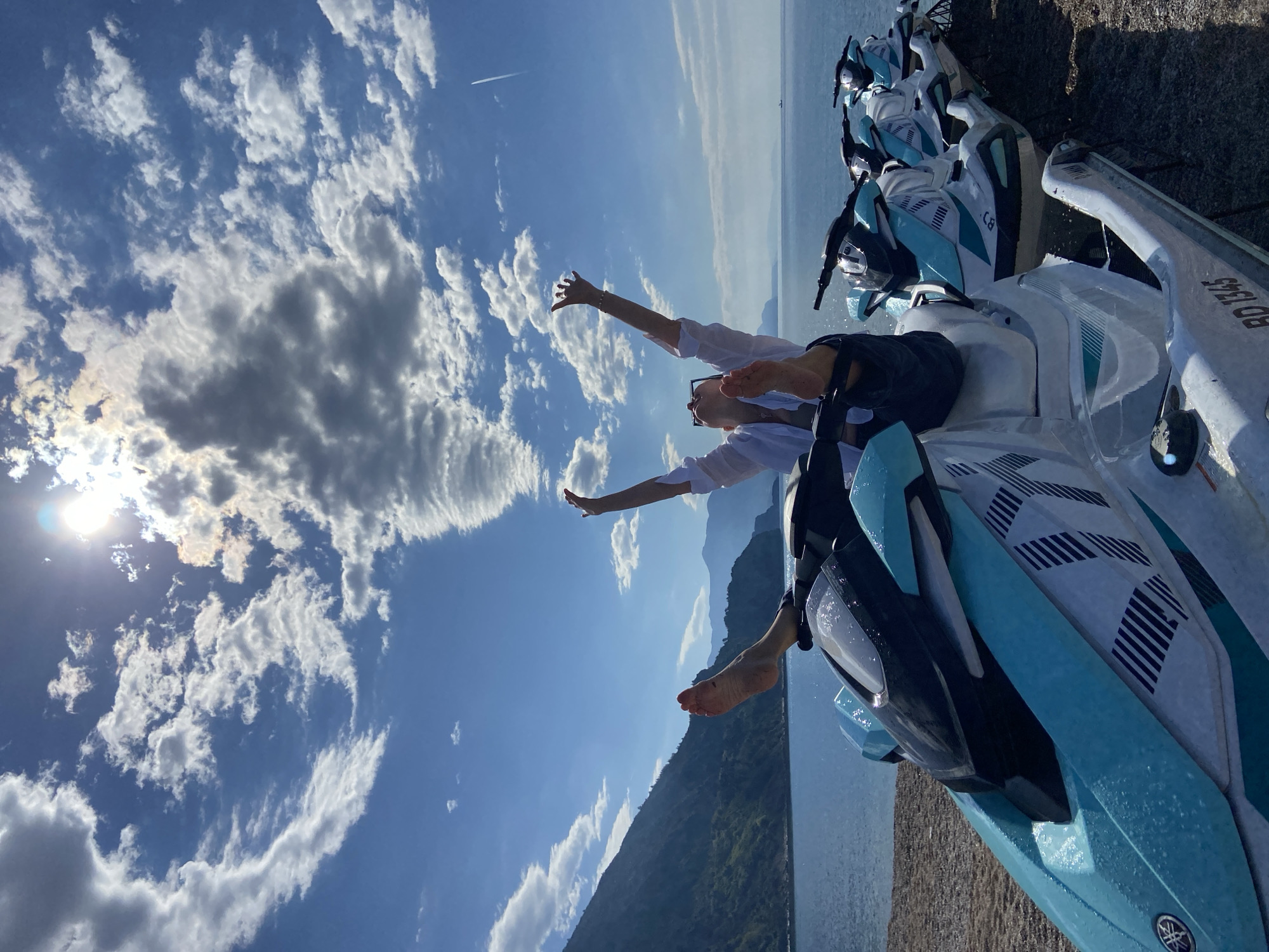 The image depicts a person lying on a blue and white jet ski. The person is wearing a white shirt and dark shorts, with their arms raised towards the sky. They are barefoot. The sky is blue with scattered white clouds, and the sun is shining brightly. In the background, there's a body of water (possibly a lake or sea) and mountains. The setting appears to be outdoors on a clear, sunny day. The jet ski is parked next to other similar jet skis. There is no indication that the scene is located in Prijevor, Montenegro.