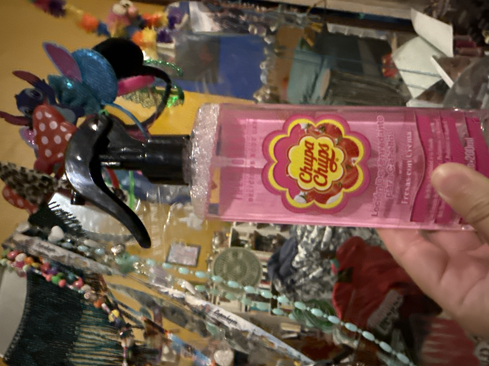 A hand holds a pink spray bottle of "Chupa Chups" brand strawberry-scented body spray in front of a cluttered scene. The scene appears to be indoors, and includes a variety of objects like headbands, necklaces, and other small trinkets. The location is not discernible from the image.