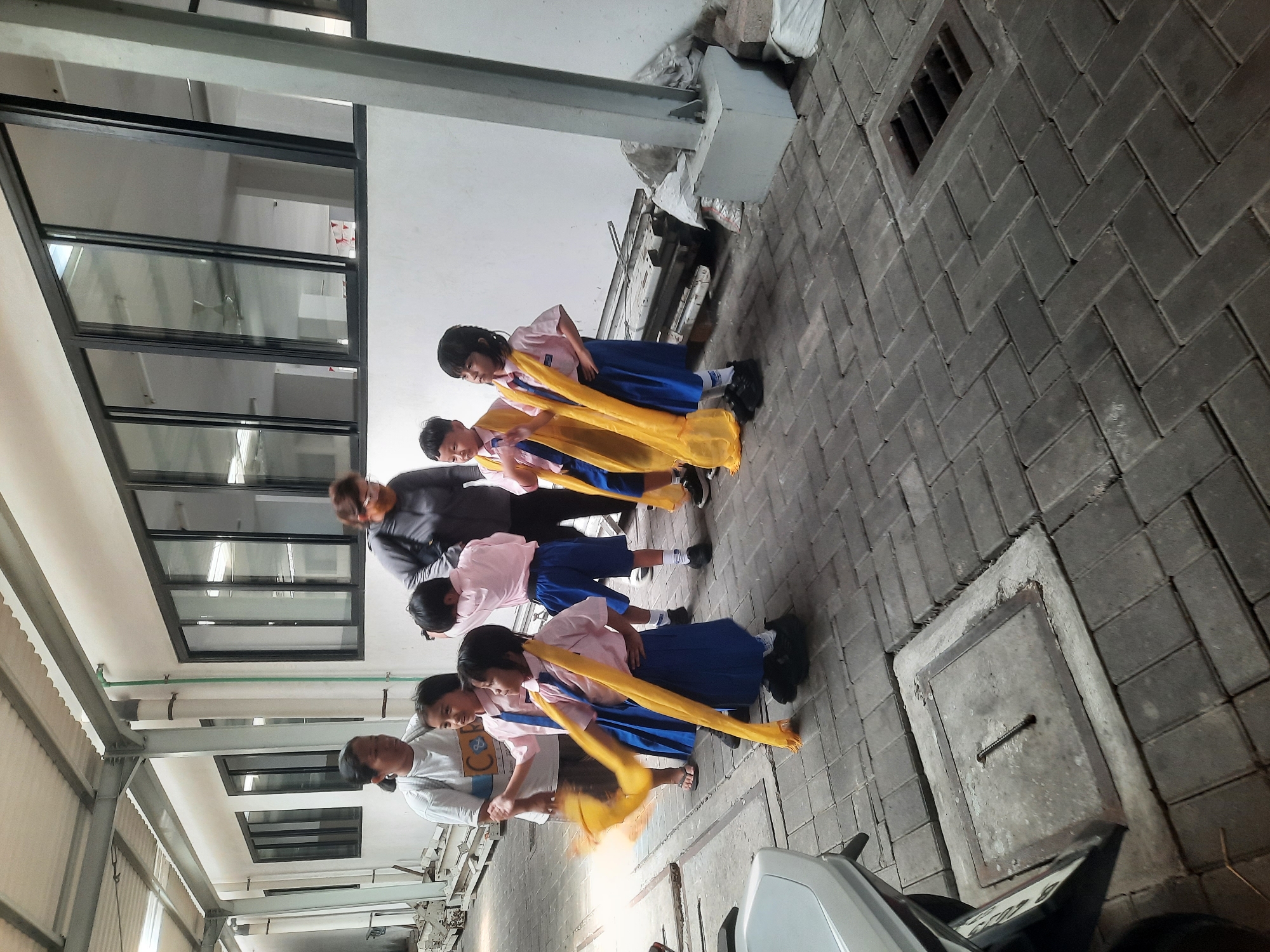 In North Jakarta, Indonesia, a group of young school children in pink shirts and blue skirts, accessorized with yellow sashes, are gathered outside a building with large glass-paneled windows. One child in the foreground is actively twirling their yellow sash. An adult woman, likely a teacher or guardian, stands to the left, observing the children. Another adult, wearing a grey jacket and a mask, is positioned behind the children. The scene is set on a paved outdoor area with interlocking grey bricks. Several metal structures and white bags are visible in the background, suggesting a construction or storage area. A drainage grate and a covered manhole are present on the ground, indicating an urban environment.