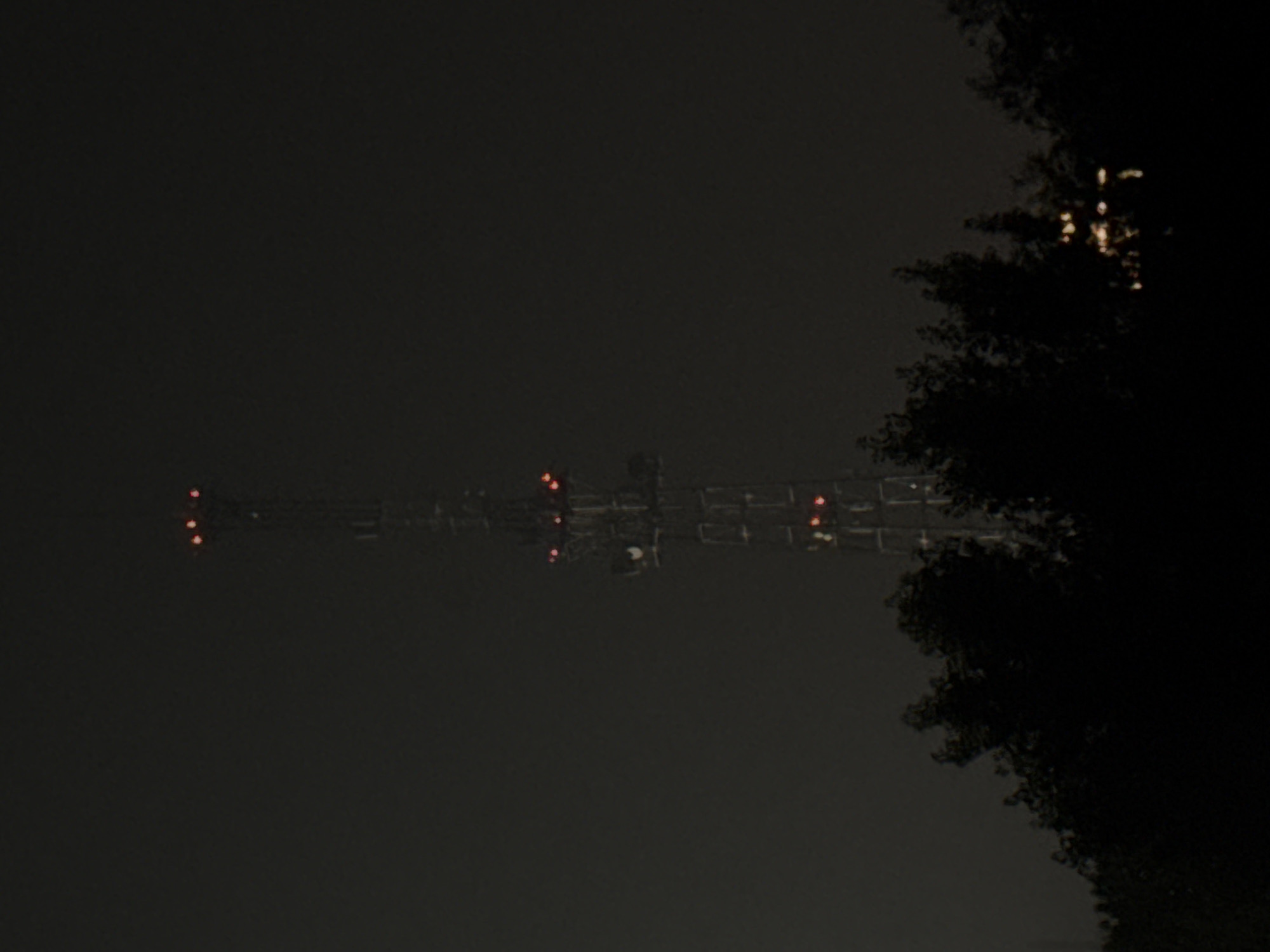 A tall, lattice-like telecommunications tower stands silhouetted against a dark, hazy sky. The tower is illuminated by several red lights near its peak and at various levels along its structure. In the foreground, a dense line of dark trees and foliage obscures the lower portion of the image, suggesting a nighttime scene with limited visibility. Some faint, yellow lights are visible through the trees in the far distance, possibly indicating a city or other populated area. The overall impression is one of a remote or industrial area at night, with the tower serving as a prominent landmark. The location is identified as Oskemen, Kazakhstan.