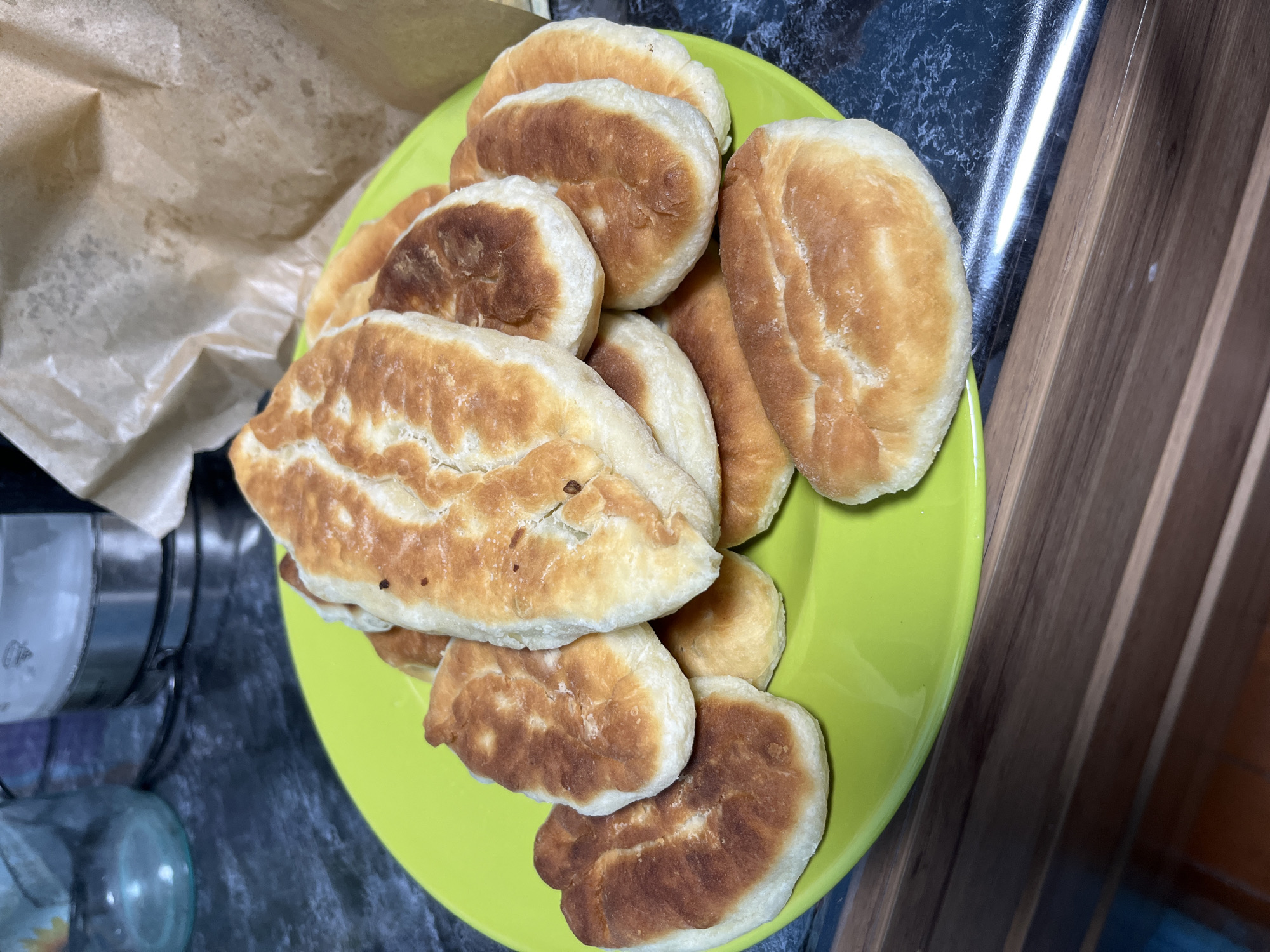 A pile of freshly fried pastries, golden brown and puffed, are arranged on a bright green plate. The pastries, which appear to be of Slavic origin, possibly pirozhki or similar, are oval-shaped with a slightly folded edge, suggesting a filling is enclosed within. Some exhibit a slightly darker, crispier crust, while others are a lighter golden hue. The plate is set on a dark, speckled countertop that reflects light. In the background, partially visible elements include a glass jar, a kettle, and a crumpled brown paper bag. The overall scene suggests a home kitchen environment, likely in Nizhny Tagil, Russia, given the context.