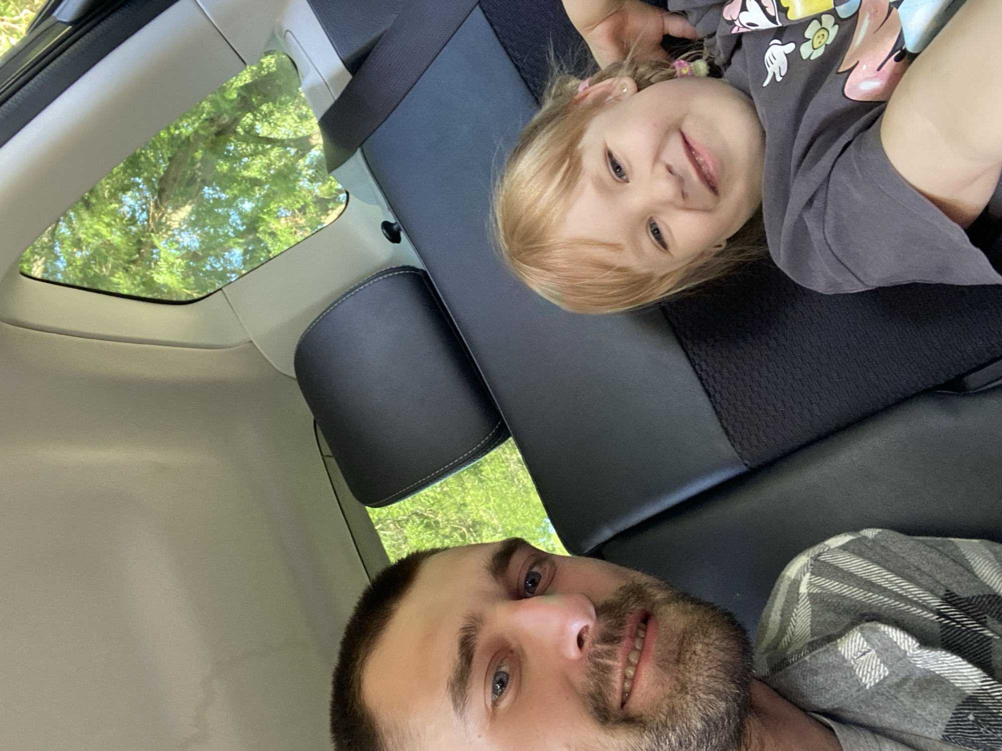 In this selfie taken from a car, a man and a young girl are the subjects, lying down on the back seats. The man, in the foreground, has short brown hair, a beard, and is smiling at the camera. He's wearing a grey and white plaid shirt over a grey t-shirt. The girl, positioned above him, has blonde hair with pink accessories, blue eyes, and is also smiling, looking towards the camera. She is wearing a dark grey t-shirt with cartoon characters on it.
The background shows the interior of a car with black leather seats and a small window through which green trees and blue sky are visible, suggesting an outdoor setting. The overall mood appears relaxed and happy, possibly during a car ride or outing. The image is taken in daylight, likely mid-day given the bright lighting filtering through the trees. The provided location context is Odesa, Ukraine. There is no visible text in the image.