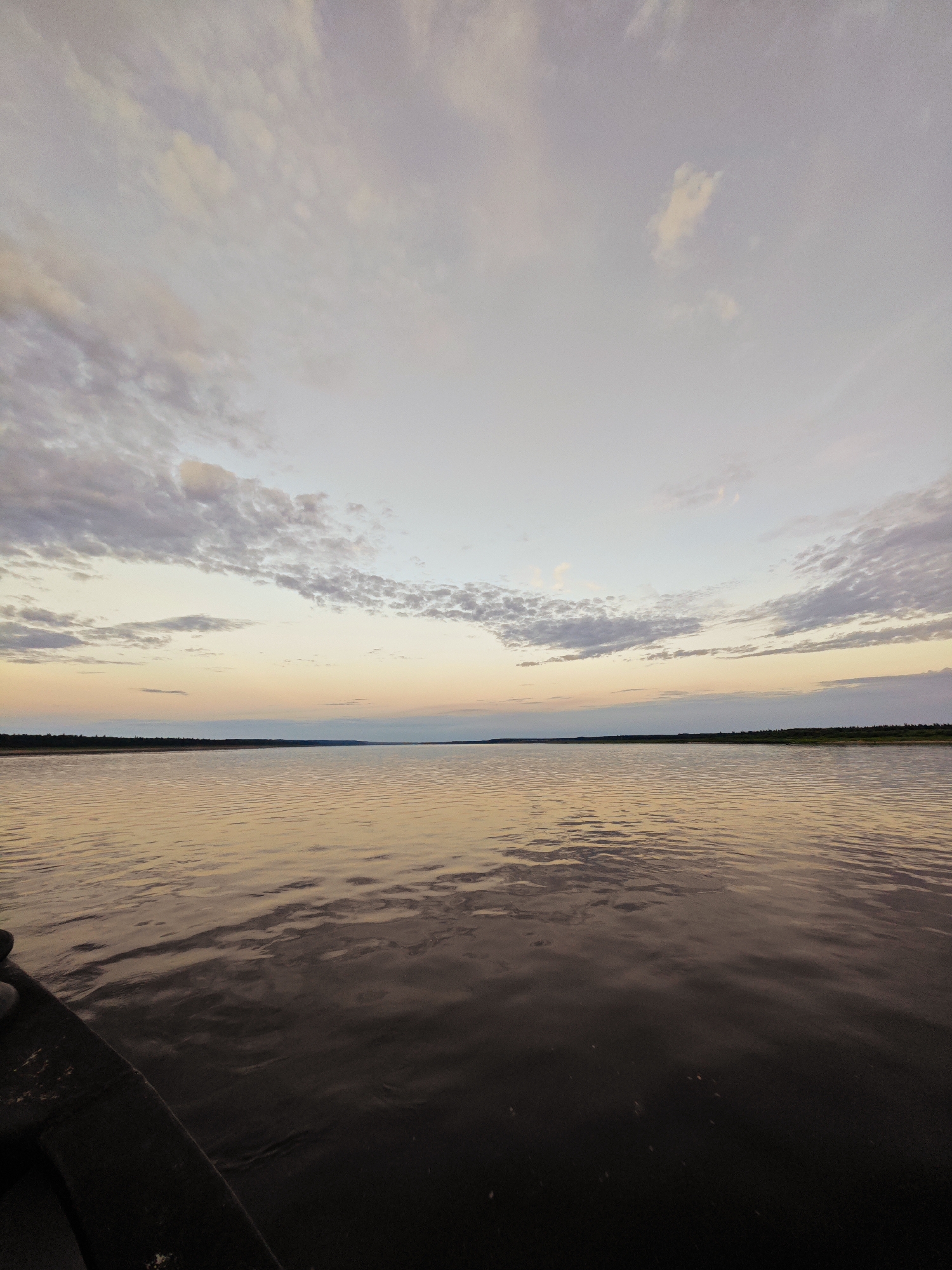 A wide shot captures a serene lake at dusk, viewed from a boat. The water, reflecting the sky's warm hues, exhibits gentle ripples. In the distance, a tree line marks the horizon, with a faint stretch of land visible. The sky is a canvas of soft pastels, transitioning from pale blue at the top to shades of pink, orange, and yellow closer to the horizon, adorned with scattered, wispy clouds. The foreground is dominated by the dark, textured edge of the boat, implying movement or a pause on the water. The overall mood is tranquil and peaceful, characteristic of a sunset over a calm body of water. There is no visible text in the image.