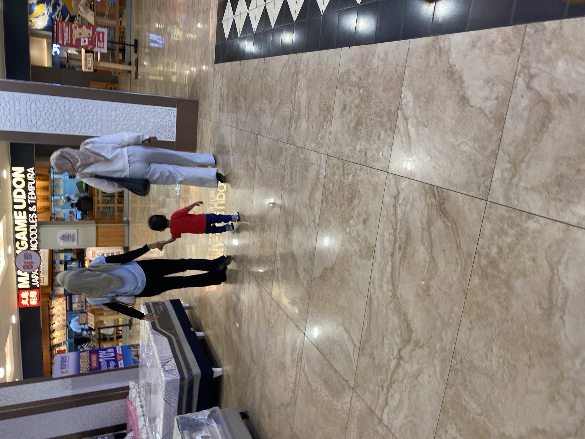 In a well-lit indoor shopping mall, a woman wearing a light blue shirt and black pants, with a beige hijab, walks hand-in-hand with a young child in a red shirt and blue pants. Following closely behind them is another woman dressed in a white outfit, including a white hijab, carrying a black tote bag. The floor is tiled with large, beige, marble-patterned tiles, and a section of black and white patterned tiles is visible on the right side of the frame. In the background, a restaurant with a prominent sign displaying "MAME GAME UDON" is visible, along with other shop displays. The environment suggests a commercial setting within a city, likely in a location with a significant Muslim population due to the attire of the individuals. While the image does not explicitly confirm the location as Banda Aceh, Indonesia, the presence of Halal signage on restaurant fronts is common in such areas.