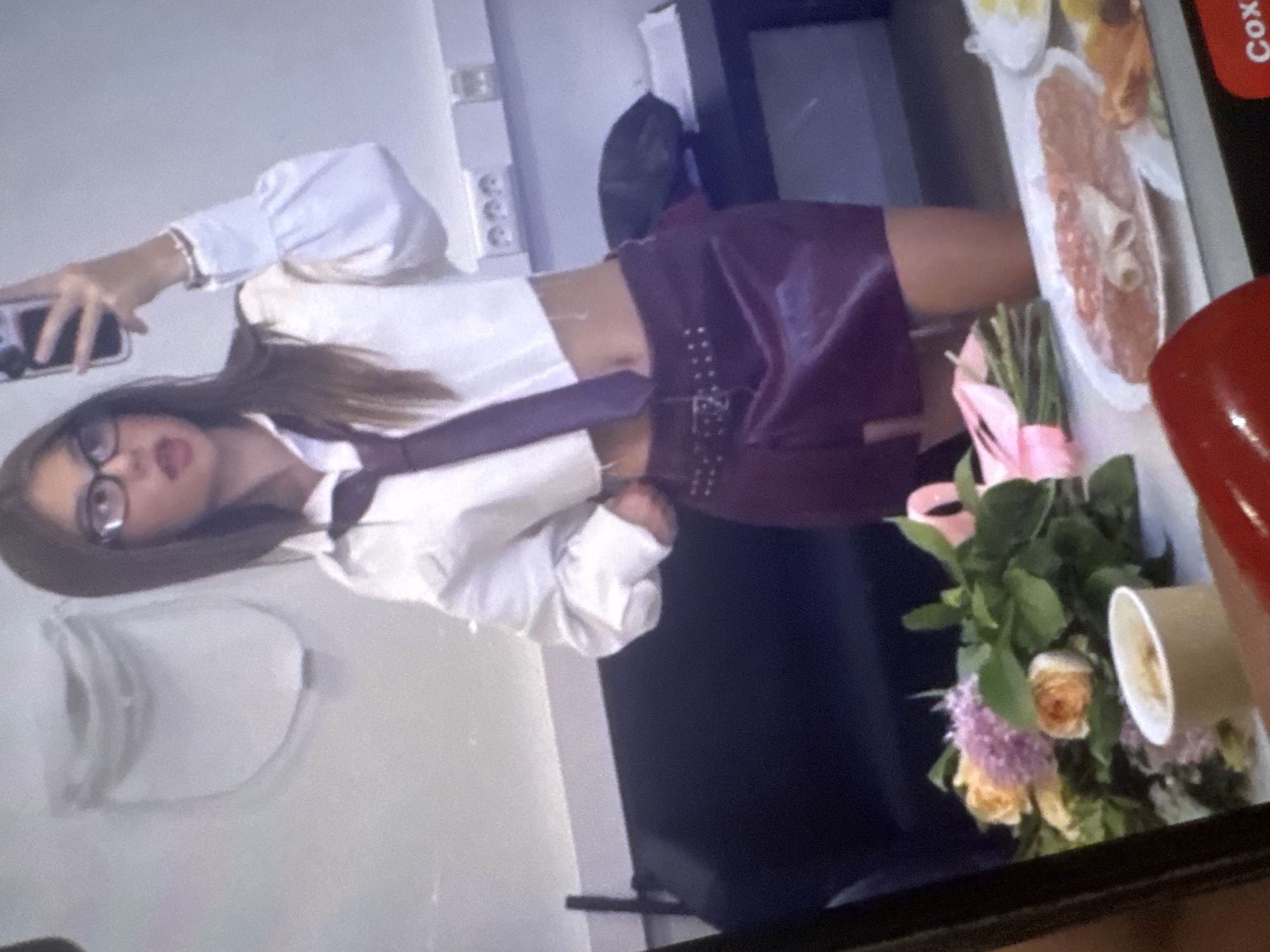 This image is a photograph of a screen, displaying a picture of a young woman in an indoor setting. The image is rotated 90 degrees counter-clockwise.
The woman, with long dark hair, is wearing glasses, a white long-sleeved shirt with puffed sleeves, and a dark tie. Her midriff is visible above a dark, possibly purple or maroon, skirt or shorts adorned with metallic studs. She holds a black-cased smartphone in her left hand, appearing to be taking a photo of herself, possibly a mirror selfie. A light-colored bracelet is visible on her left wrist. Her mouth is slightly open, and she looks towards the camera.
To the right of the woman (as she would be oriented upright), a rectangular platter of food is visible, featuring sliced meats (possibly cold cuts or salami) and other yellowish items. Below the food platter, a bouquet of flowers is arranged, including yellow or peach roses, purple clusters of flowers, and green foliage, tied with a pink ribbon. A white paper cup stands next to the flowers.
The background primarily consists of a light-colored wall or surface, with what appears to be an electrical outlet. The image quality suggests it is a photo of a digital screen, with some graininess. A small portion of a red object, possibly a fingernail or part of a phone case, is visible in the bottom right corner, indicating another person is holding the camera that captured this image of the screen. Partially visible text "Cox" on a red background appears along the right edge of the displayed screen.