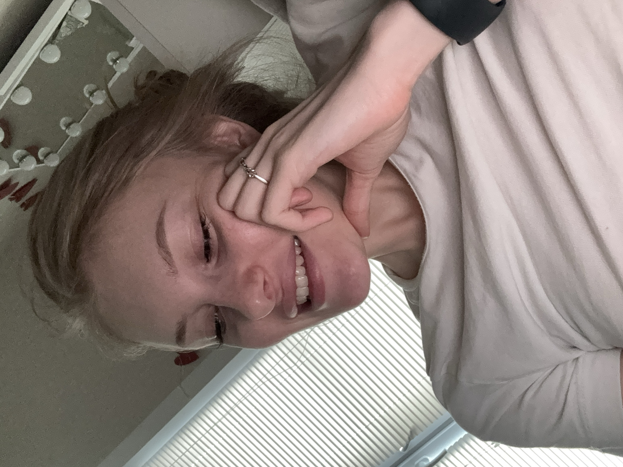 The media file is a close-up, low-angle, portrait-style photograph of a young woman. She is looking towards the viewer with a smile, her head tilted and resting on her hand, which is supporting her face. Her eyes are open, and her teeth are visible in her smile. She has light-colored hair pulled back. She is wearing a light-colored t-shirt, and a black band, possibly a watch or fitness tracker, is visible on her wrist. A ring is on her right index finger.
The background of the image includes a mirror with several light bulbs, suggesting a vanity or dressing area. Below the woman, there are horizontal blinds, indicating a window. The overall lighting suggests it could be daytime, possibly indoors. The subject's expression conveys happiness and perhaps a sense of playfulness or tiredness. The image has a slightly blurry quality. No visible text is present in the image.