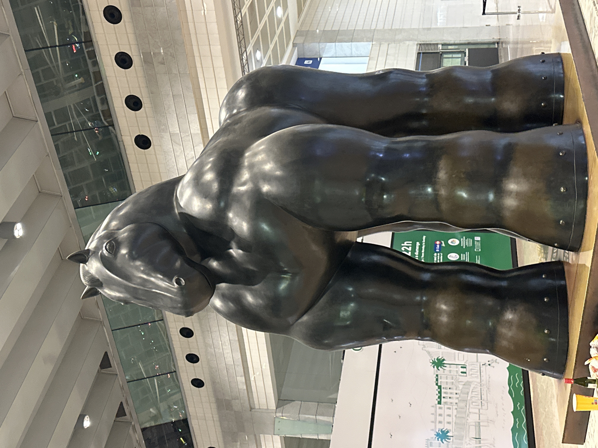 The image shows a large, dark bronze sculpture of a horse with an exaggeratedly muscular physique, standing indoors. The sculpture is positioned on a polished floor, likely marble or granite, with a slight sheen reflecting the overhead lighting. Behind the sculpture, a large white wall features a green and white mural depicting a coastal scene with palm trees and buildings. To the right of the mural, a green sign with white text, likely informational, is partially visible. The background reveals an indoor architectural setting with a high ceiling, exposed metal beams, and large windows reflecting lights. The sculpture is the primary subject, dominating the frame with its imposing presence. There are no visible people interacting with the sculpture. In the foreground, to the left, a yellow cup and a small bottle of wine are placed on the floor. The overall environment suggests a public space, potentially a museum, airport, or convention center.
The sculpture of the horse, titled "The Horse" or "El Caballo" by Fernando Botero, is located in the arrivals hall of Barcelona–El Prat Airport (BCN) in El Prat de Llobregat, Spain.