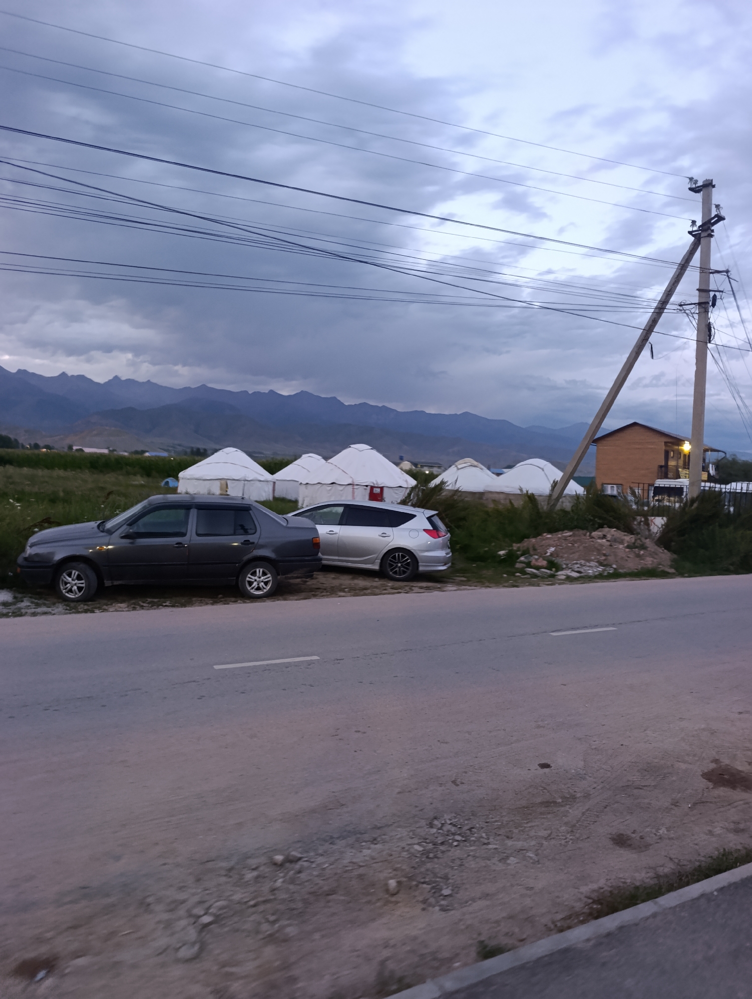 A rural roadside scene in Bosteri, Kyrgyzstan, captures a twilight setting under a cloudy sky with distant mountains. A grey sedan and a silver hatchback are parked alongside a paved road. In the background, several traditional yurts stand in a grassy field, suggesting a nomadic or semi-nomadic settlement. A wooden house with balconies is situated near a power pole with multiple wires stretching across the frame. The ground along the roadside is a mixture of dirt, gravel, and sparse vegetation, with tire tracks visible on the road's shoulder.