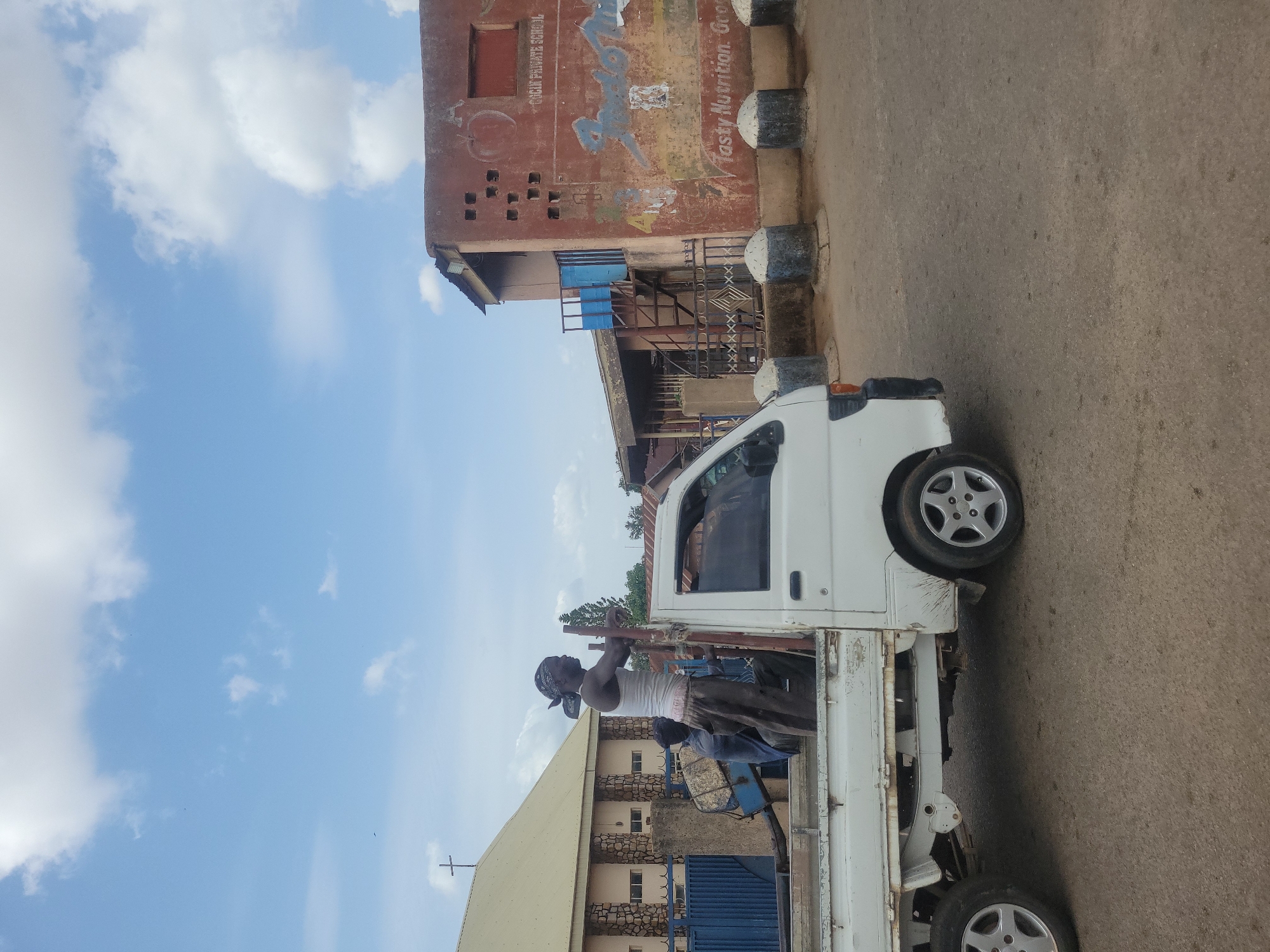 A street scene in Jos, Nigeria, features a white compact truck parked on the asphalt. A man wearing a white tank top and head covering stands in the truck bed, interacting with metal poles. In the background, buildings are visible, including a brick structure with faded signage for "Josly Private School" and "Tasty Nutrition." The sky is partially cloudy.