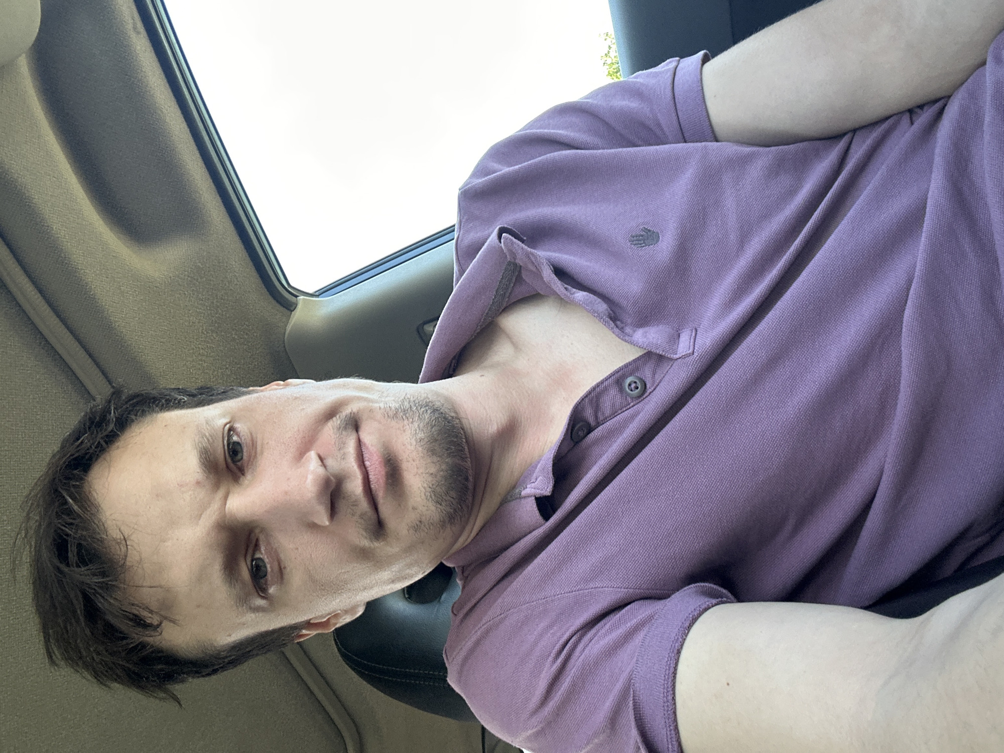 A close-up, over-the-shoulder shot captures a man seated in a car, facing the camera. He is wearing a short-sleeved, lavender polo shirt with two buttons at the collar. The man has light skin, brown hair, and stubble. The car's interior is visible, with the headliner and a portion of the passenger seat. The background outside the car window is bright and overcast. The image's metadata indicates it was taken in Makiv, Ukraine.