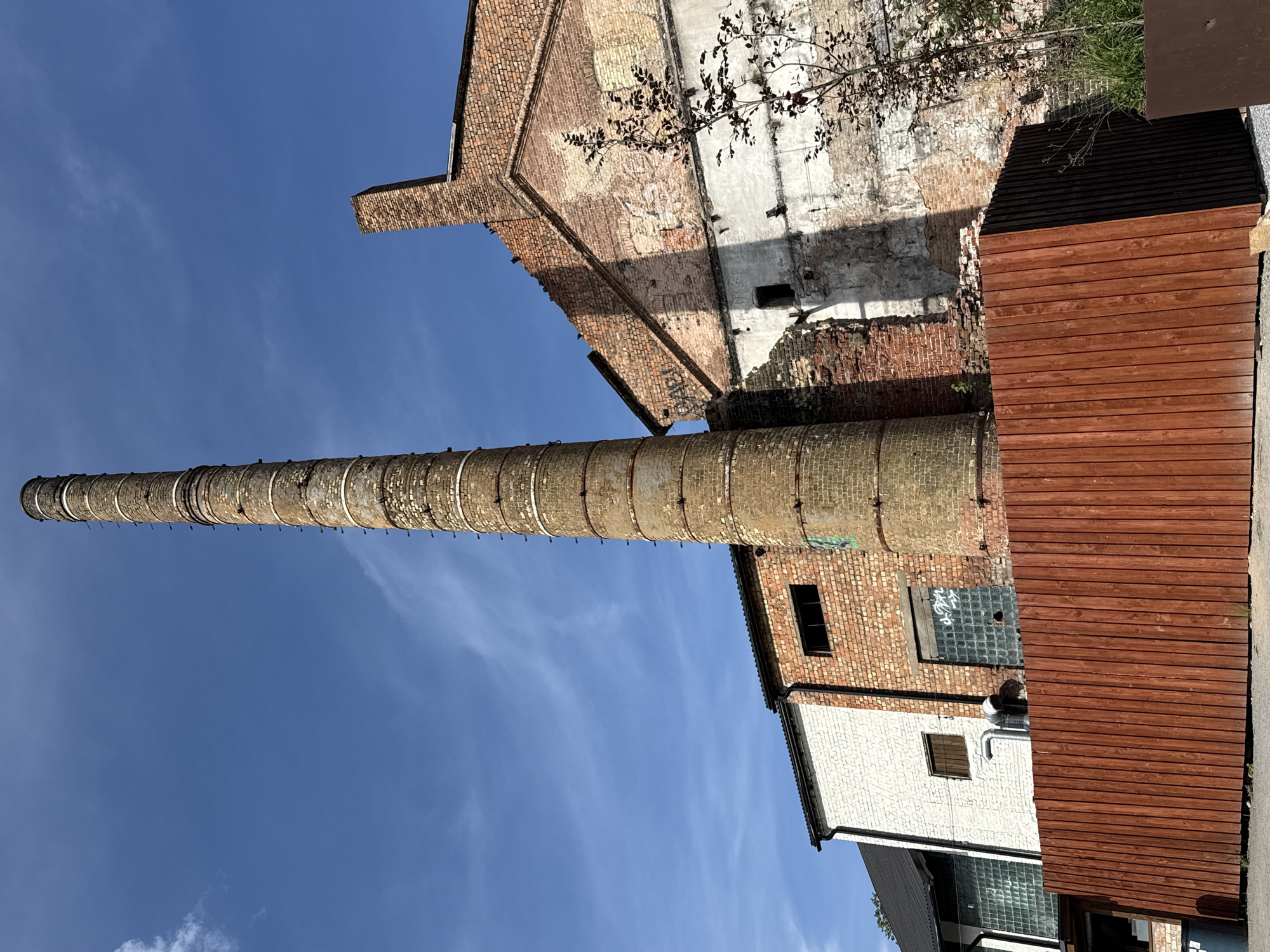 The image depicts an outdoor scene with an industrial, derelict aesthetic, likely in Riga, Latvia. Dominating the frame is a tall, cylindrical brick smokestack, weathered and showing signs of age. To its right, a crumbling brick building with exposed sections of internal structure stands under a clear blue sky. A wooden fence, reddish-brown in color, runs across the foreground, partially obscuring the base of the smokestack and the building. Behind the fence, to the left, a section of a modern building with white brickwork and glass elements is visible, contrasting with the older structures. The overall impression is one of urban decay and potential redevelopment, with elements of industrial history meeting contemporary architecture. No people are present in the image.